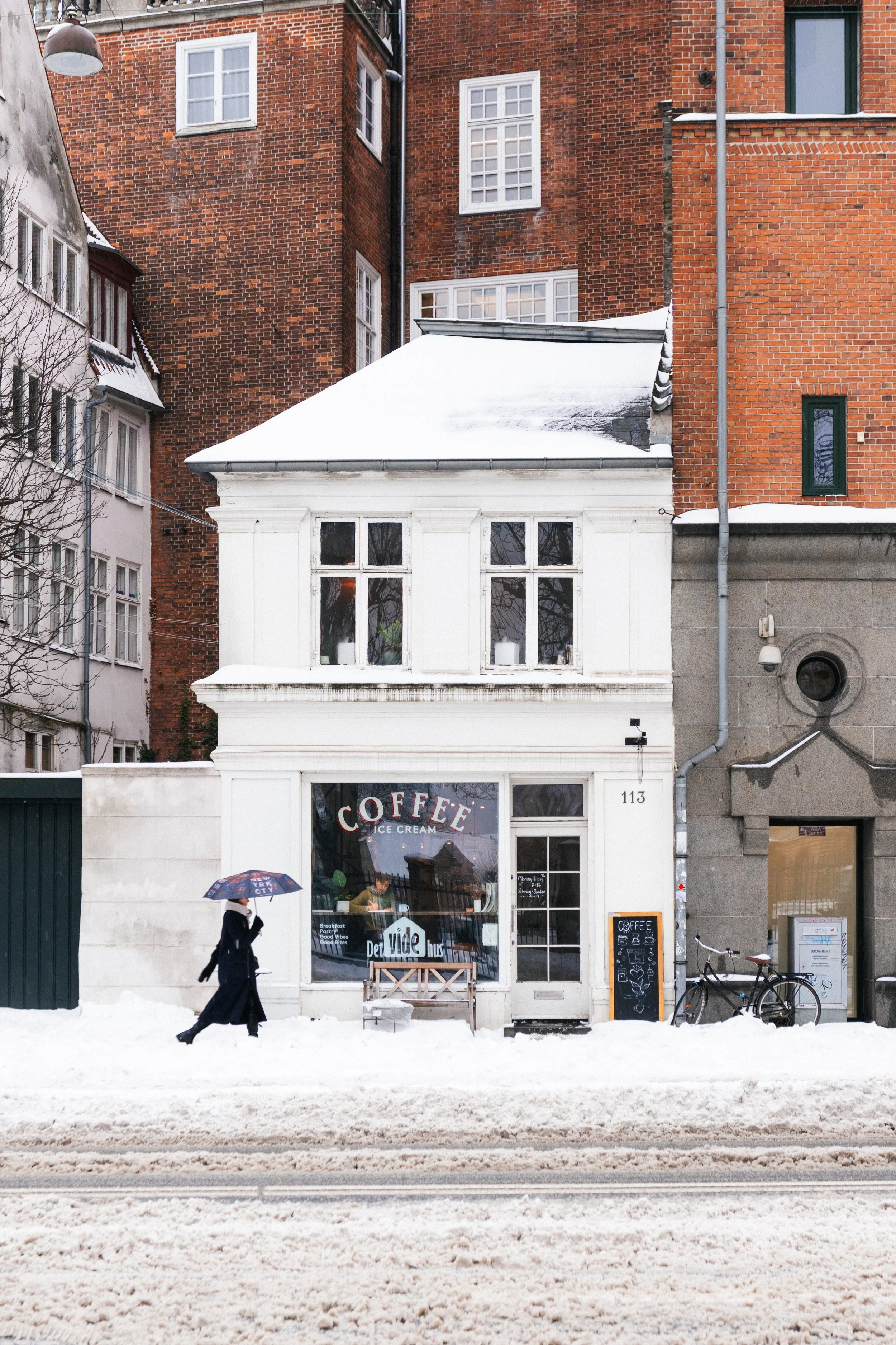 A person dressed in black walking with a colorful umbrella outside a white cafe on a snowy street. The cafe has large windows with signs advertising coffee, ice cream, and breakfast. Bicycles are parked nearby, and the scene is set in winter with sno