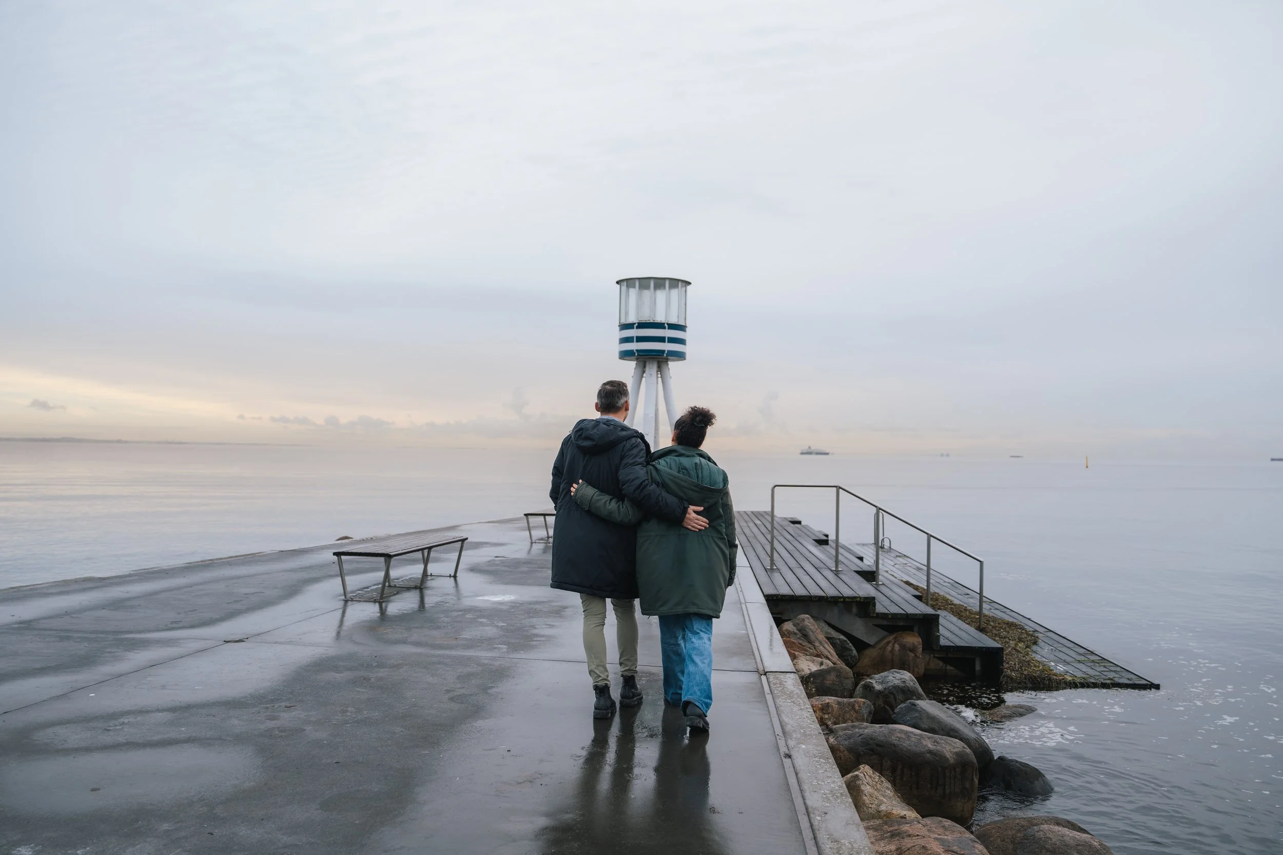 Two people walking along a waterfront pier on a cloudy day, holding each other, with a lighthouse in the background and boats in the water.