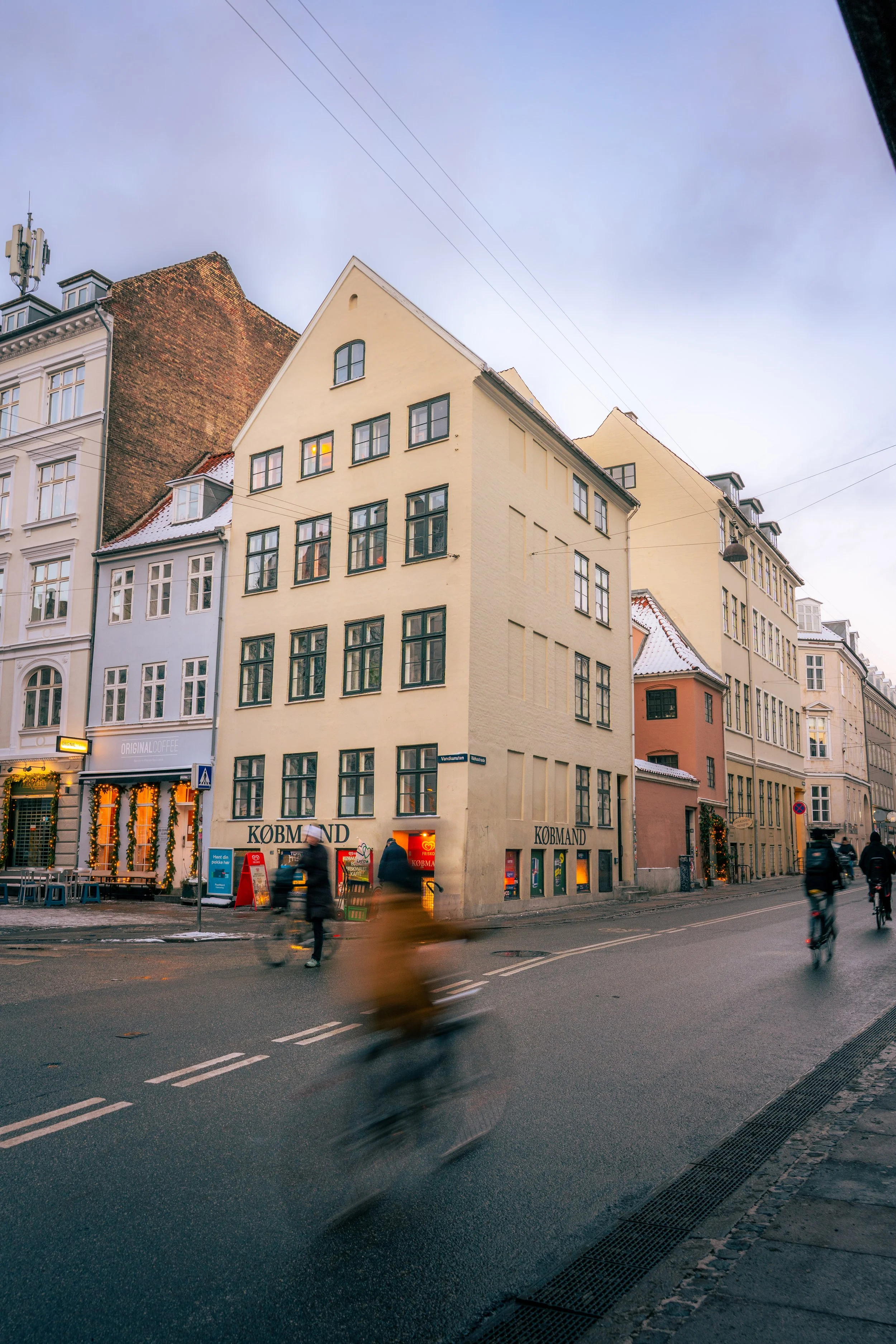 Tall beige and white buildings along a city street in the evening. Pedestrians and cyclists are moving on the street, some with motion blur, and storefronts are illuminated at the ground level.