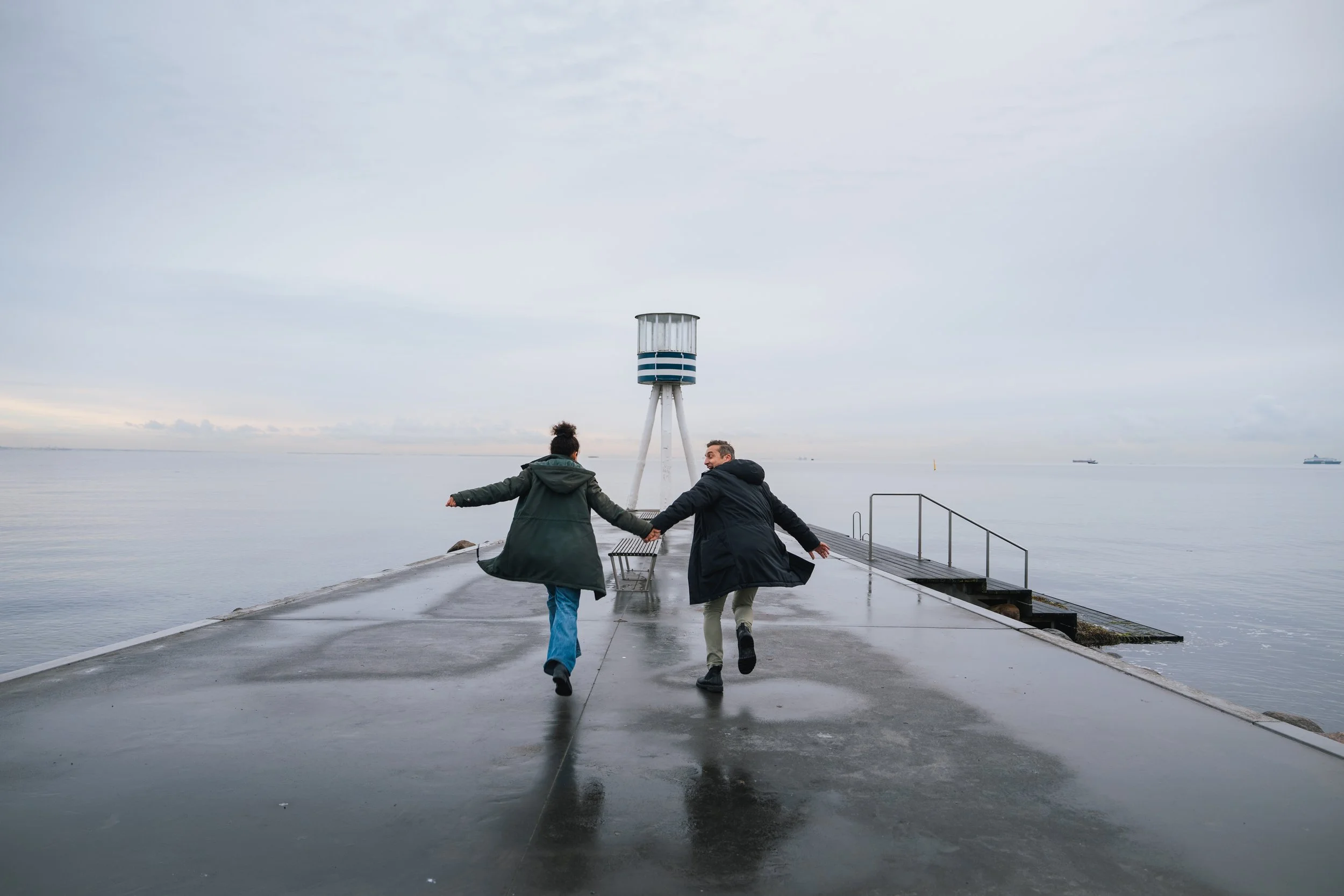 Two people holding hands and running on a wet pier near the water, overcast sky, with a lighthouse structure in the background, and ships visible on the horizon.
