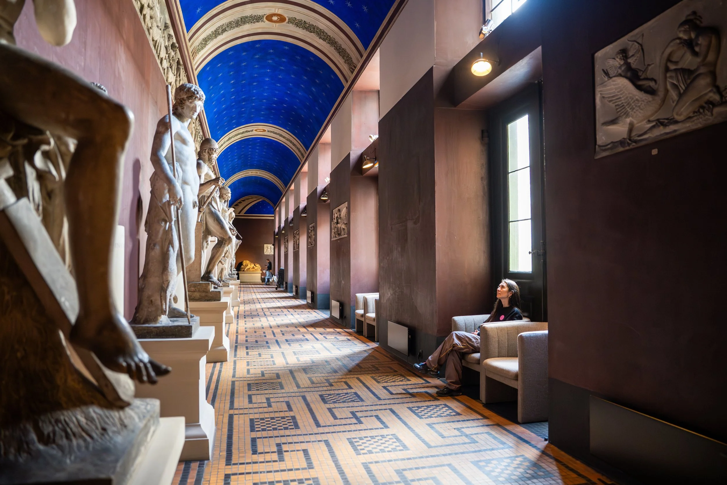Hallway with classical statues on the left and modern seating on the right, high blue vaulted ceiling with gold decorative trim, sunlight streaming through large windows on the right, and a patterned floor.