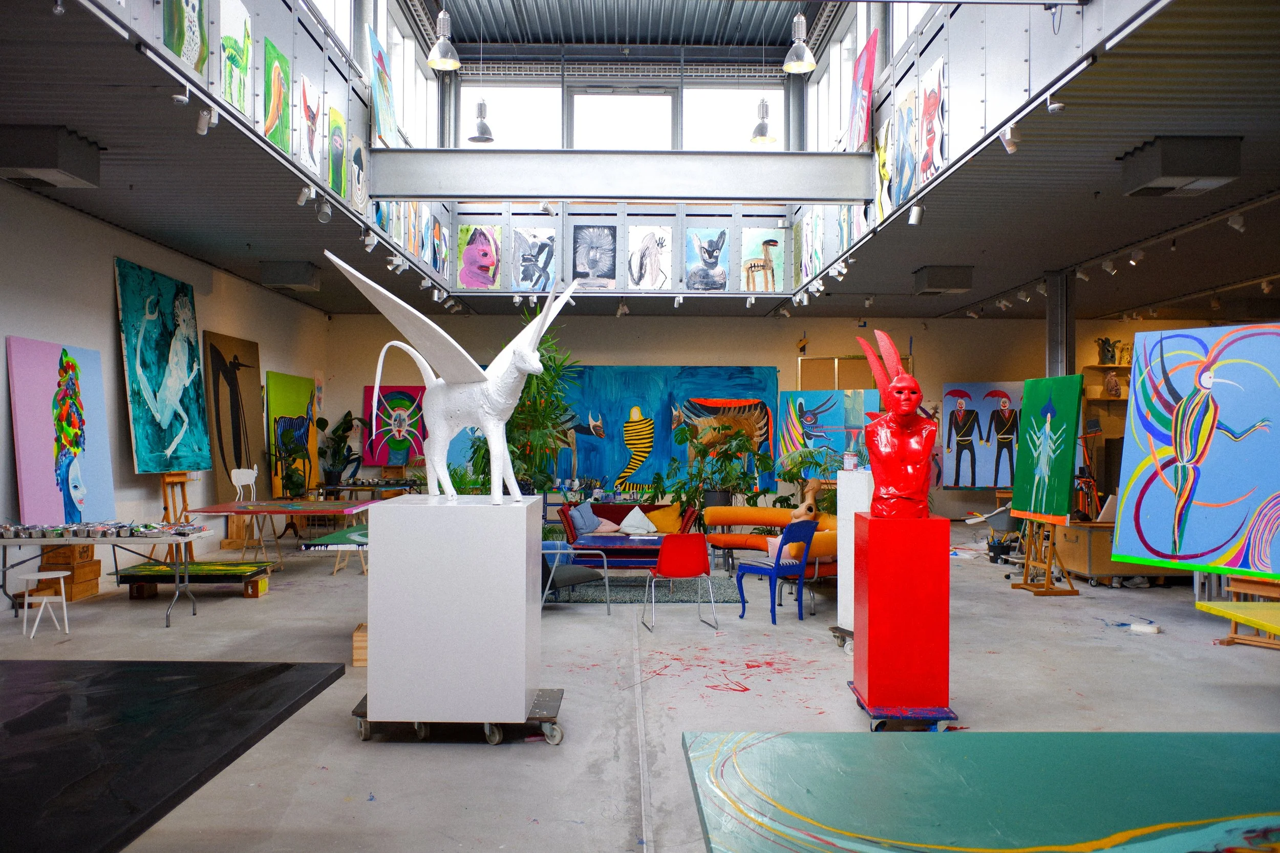 An art gallery filled with colorful paintings on walls and easels, sculptures, and furniture, with a high ceiling and bright natural light coming through large windows.