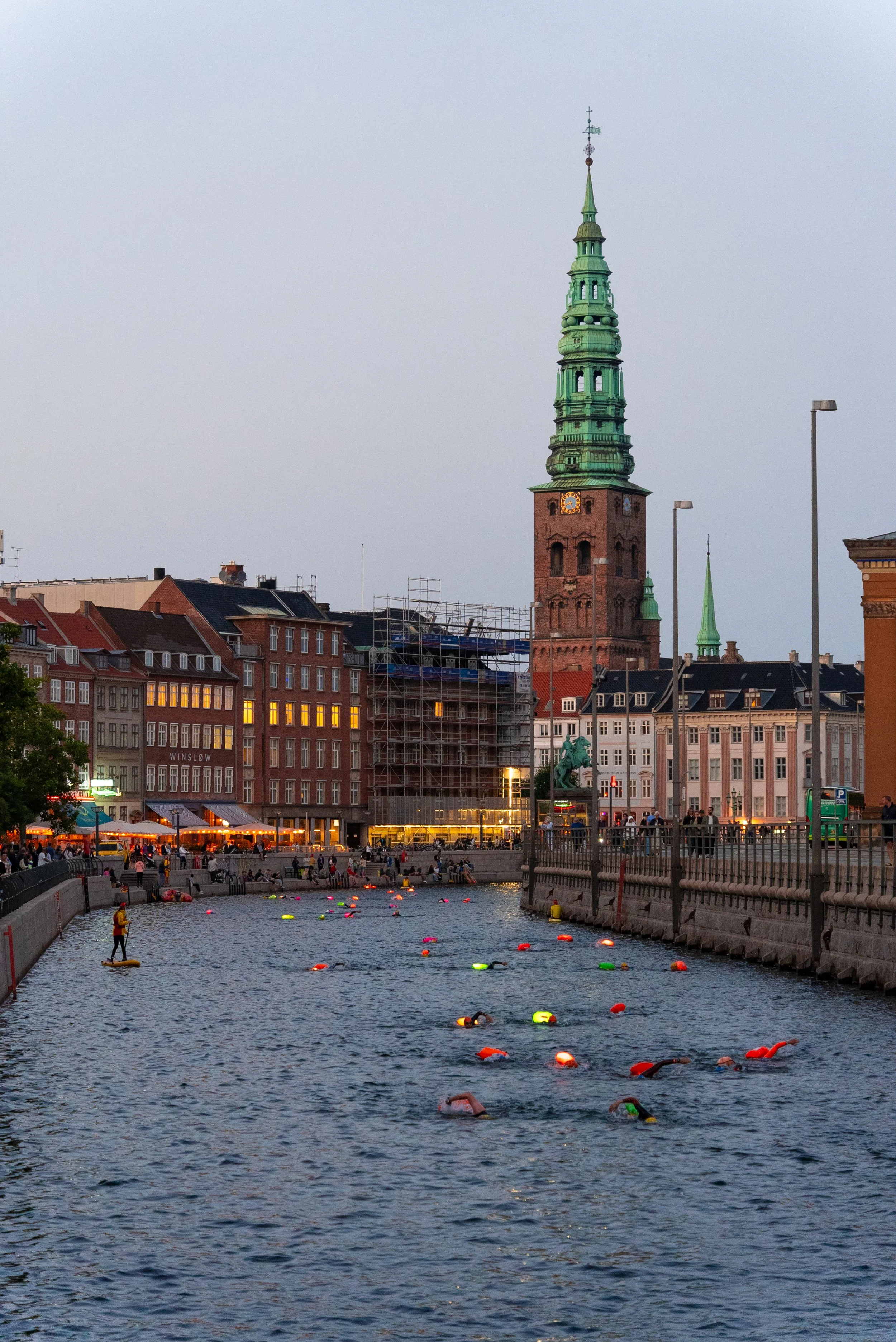 A cityscape with a tall, ornate clock tower with a green roof and spire, and a river in the foreground with people paddleboarding and floating toys, along with buildings and streetlights at dusk.