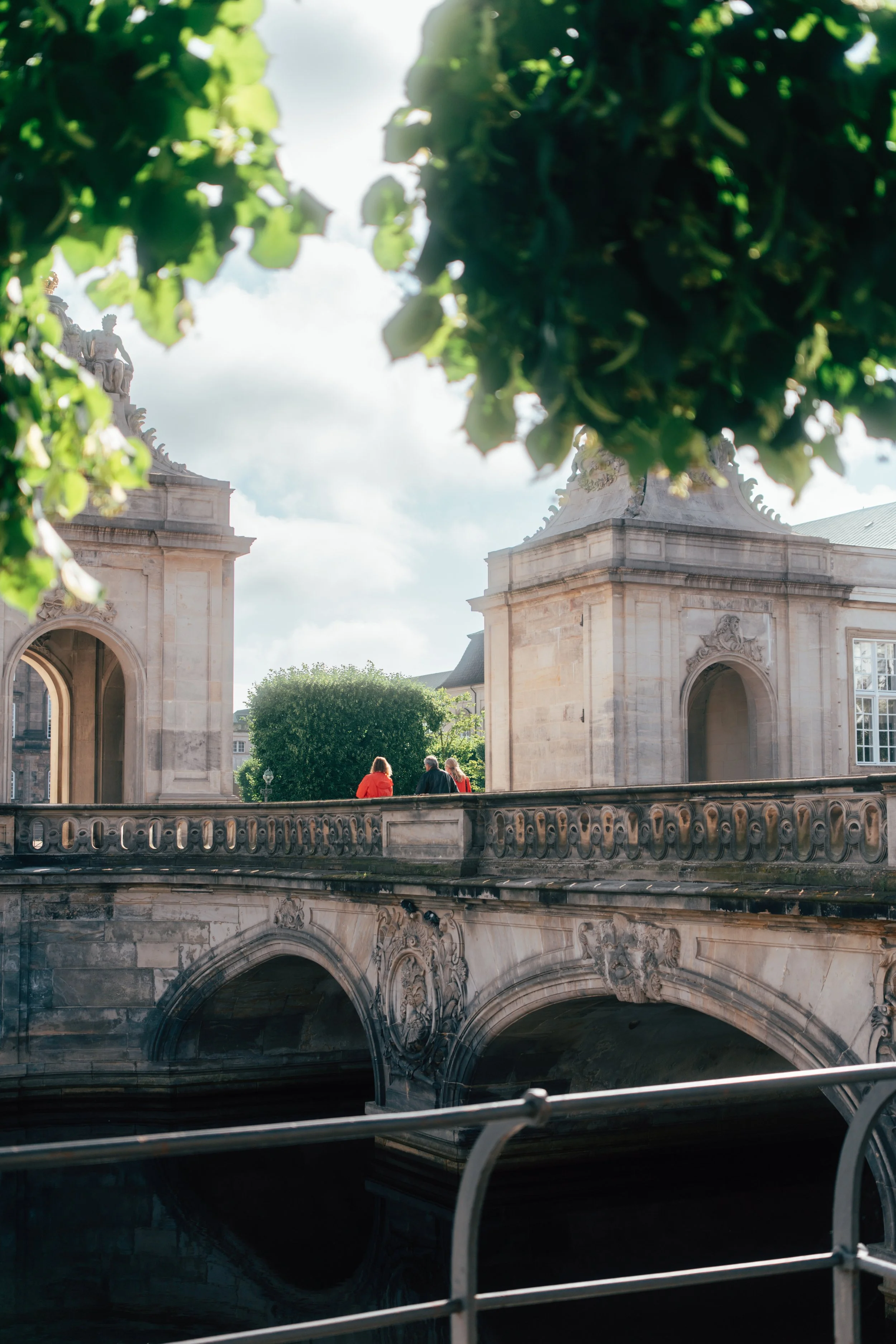 The image features the upper part of an ornate stone bridge with decorative sculptures, viewed from below through a gap framed by bright green leafy trees. The sky is partly cloudy, and three people are seen sitting on the bridge, back facing the cam