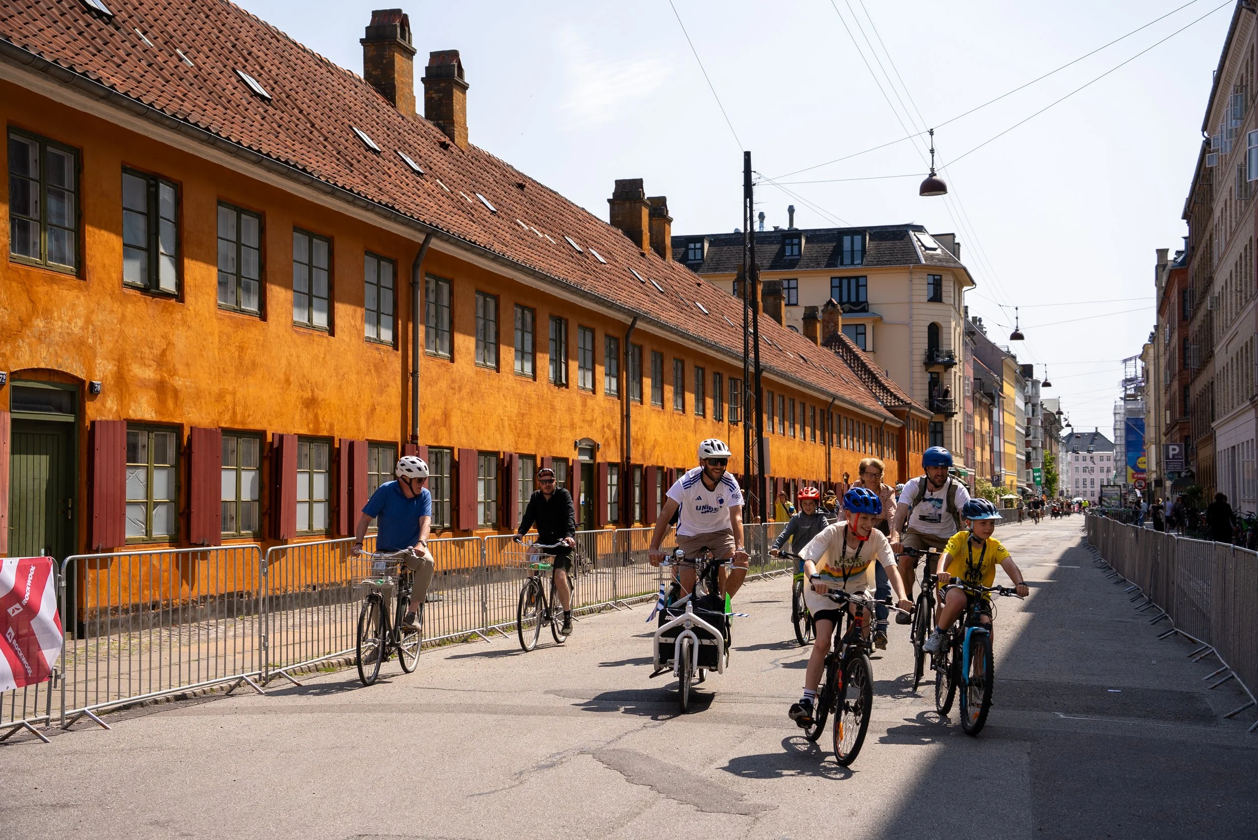 People riding bicycles and walking in an urban street with colorful buildings and overhead tram wires.