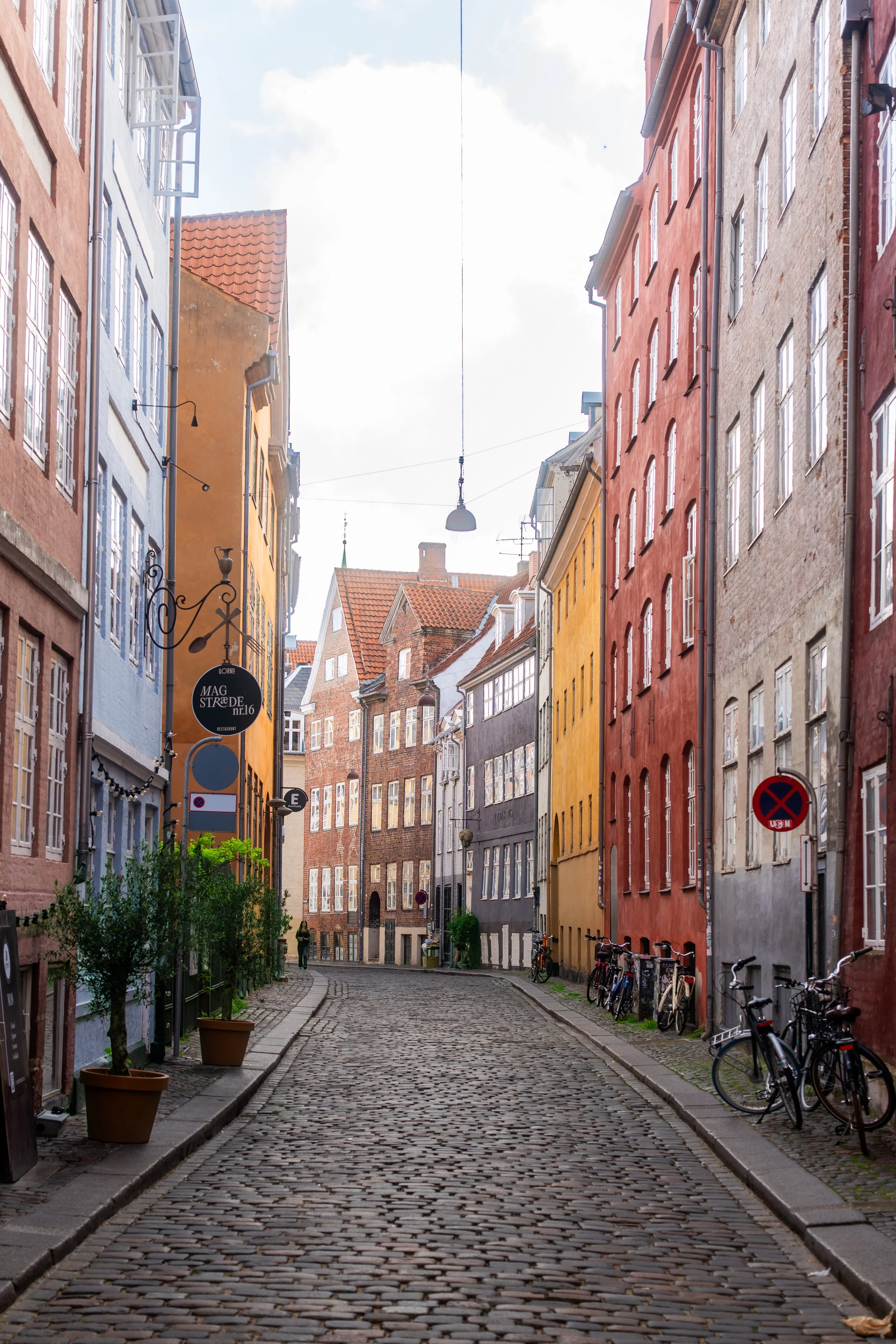 A cobblestone street with colorful buildings on both sides, some bicycles parked along the sidewalk, and a few pedestrians walking.