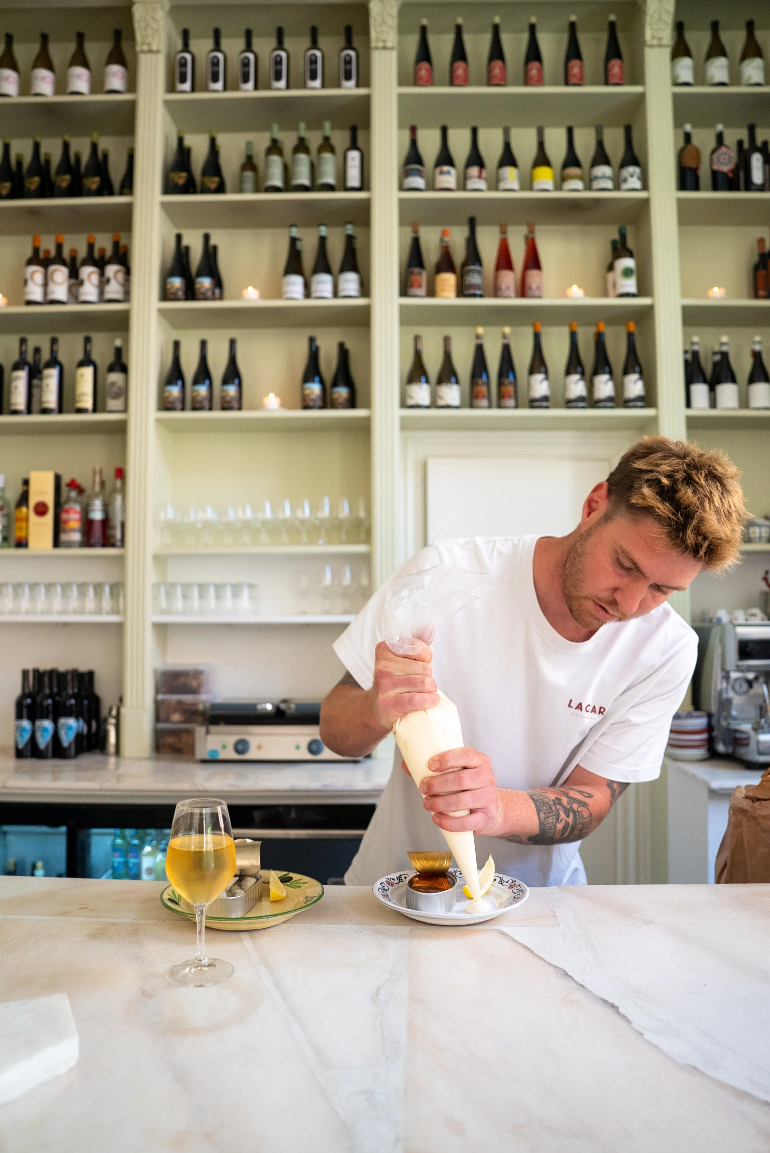 A man in a white t-shirt with tattoos on his right arm prepares a dessert in a restaurant or cafe. He is squeezing cream from a pastry bag onto a plate with caramel sauce, a small bowl, and a glass of white wine on the counter. Behind him are shelves