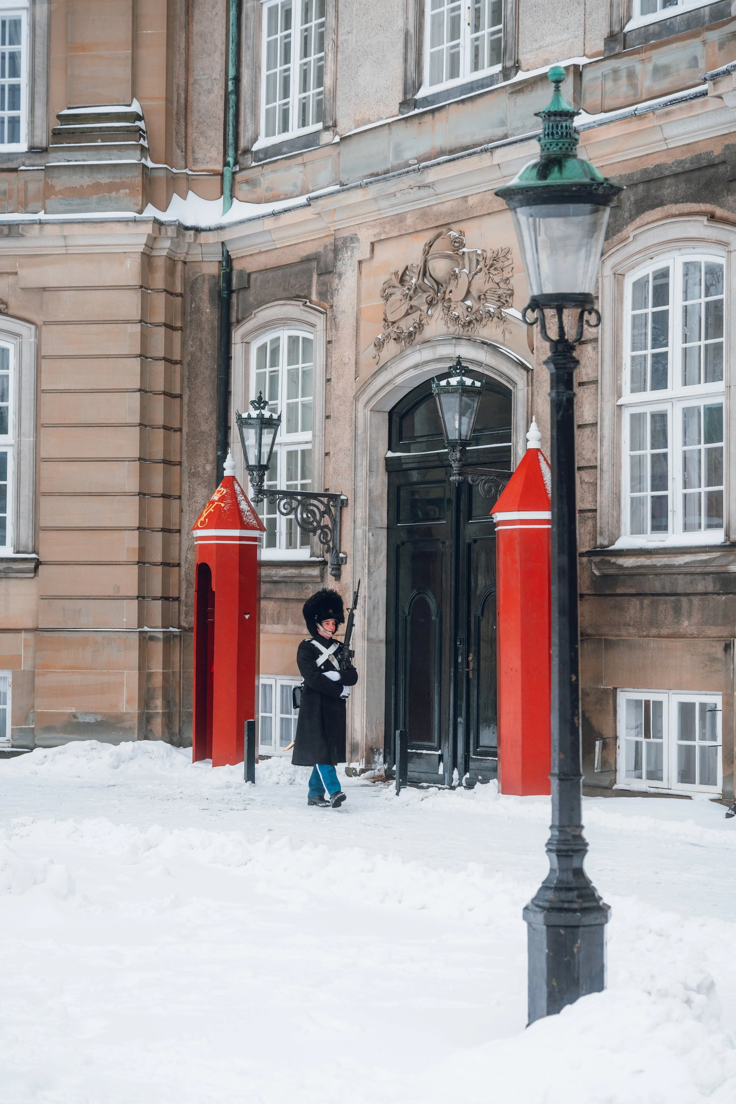 A guard post in snow outside a historic building with large windows, ornate details, and street lamps.