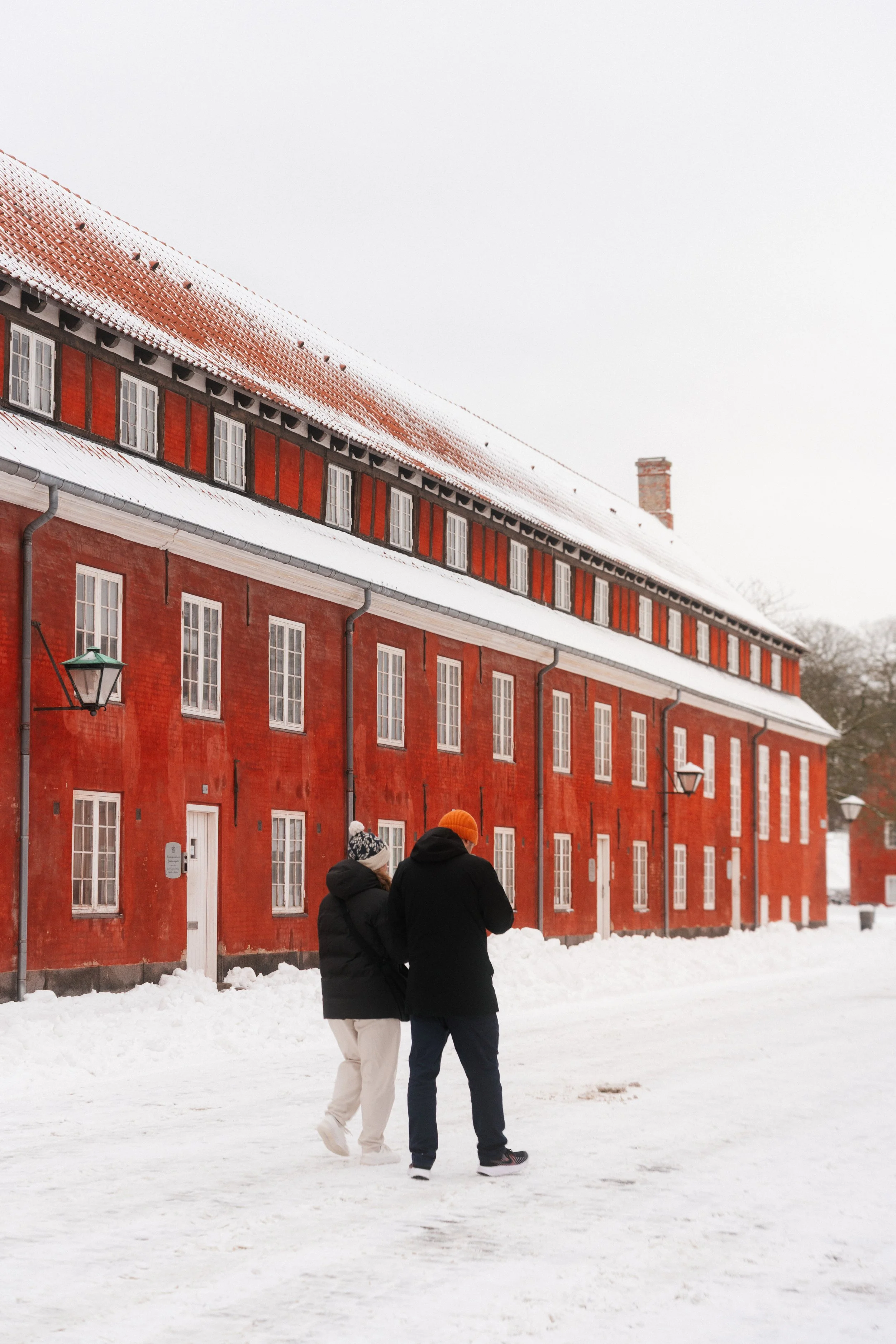 Two people walking on a snow-covered street near a large red brick building with multiple white window frames and snow on the roof, during winter.