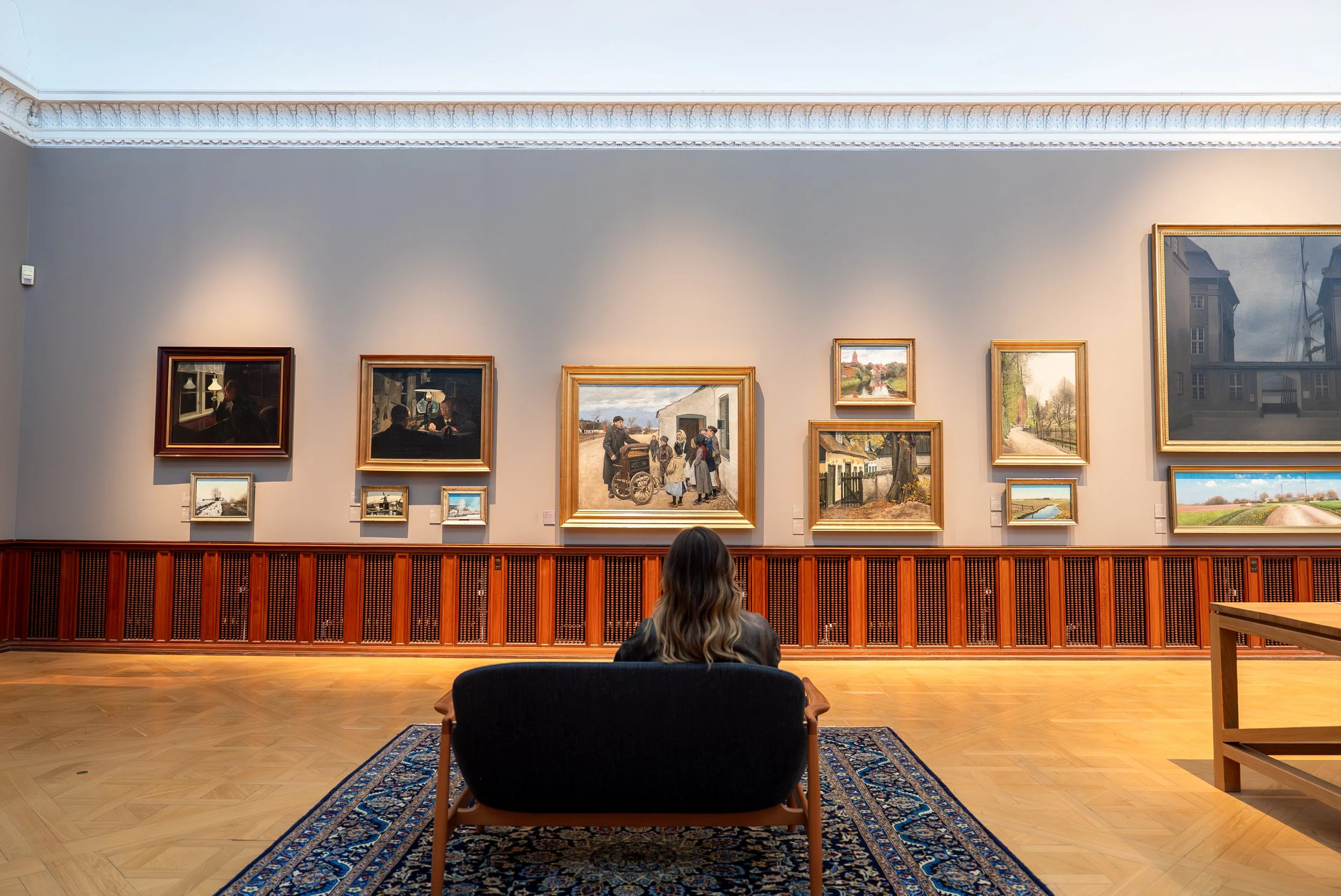 A person sitting on a chair in an art gallery, viewing framed paintings on a beige wall. The gallery has wooden flooring, a patterned rug, and decorative molding on the ceiling.