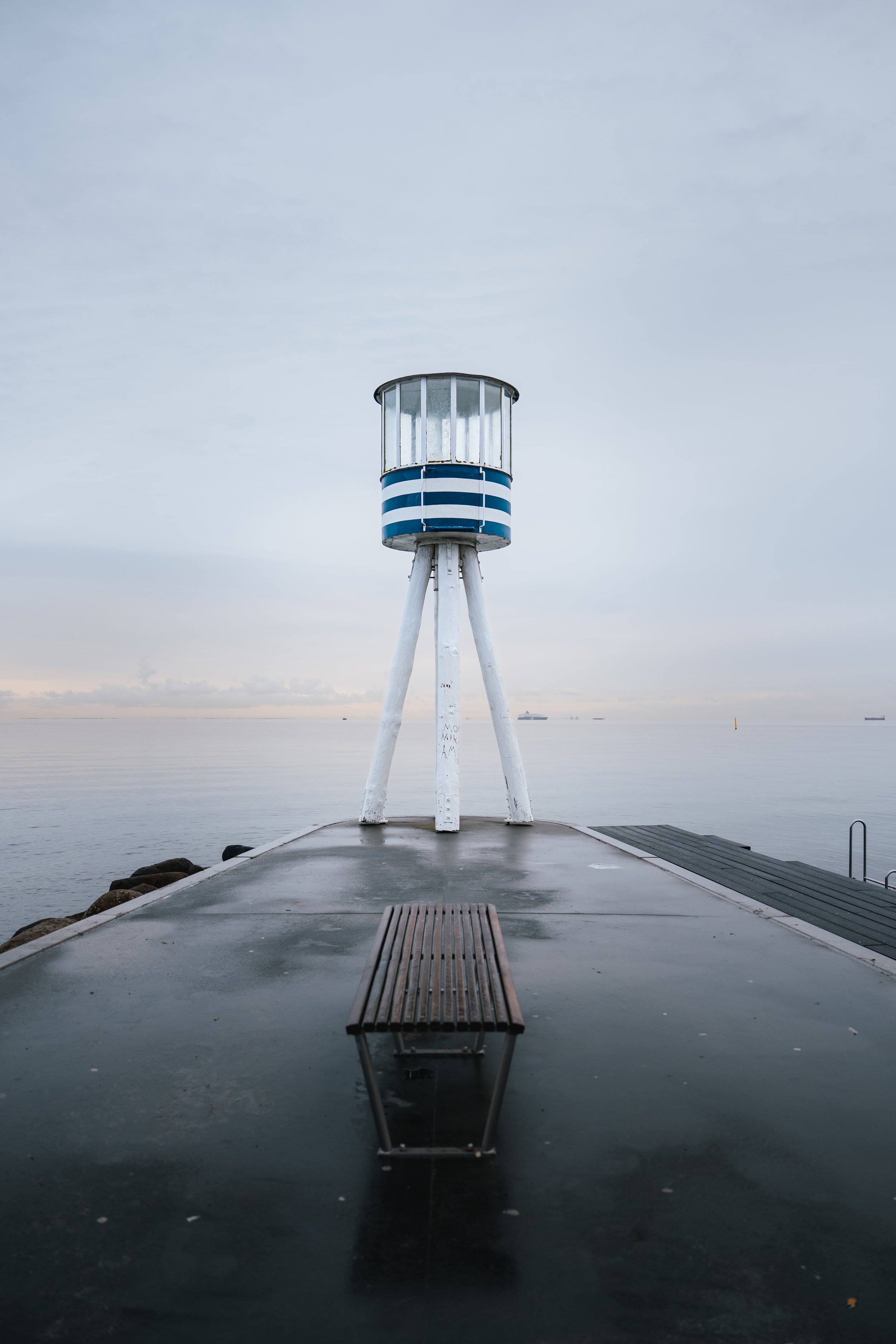 A white lighthouse with blue and white horizontal stripes sits on three white legs on a pier by the water, with a cloudy sky and distant ships visible in the background.