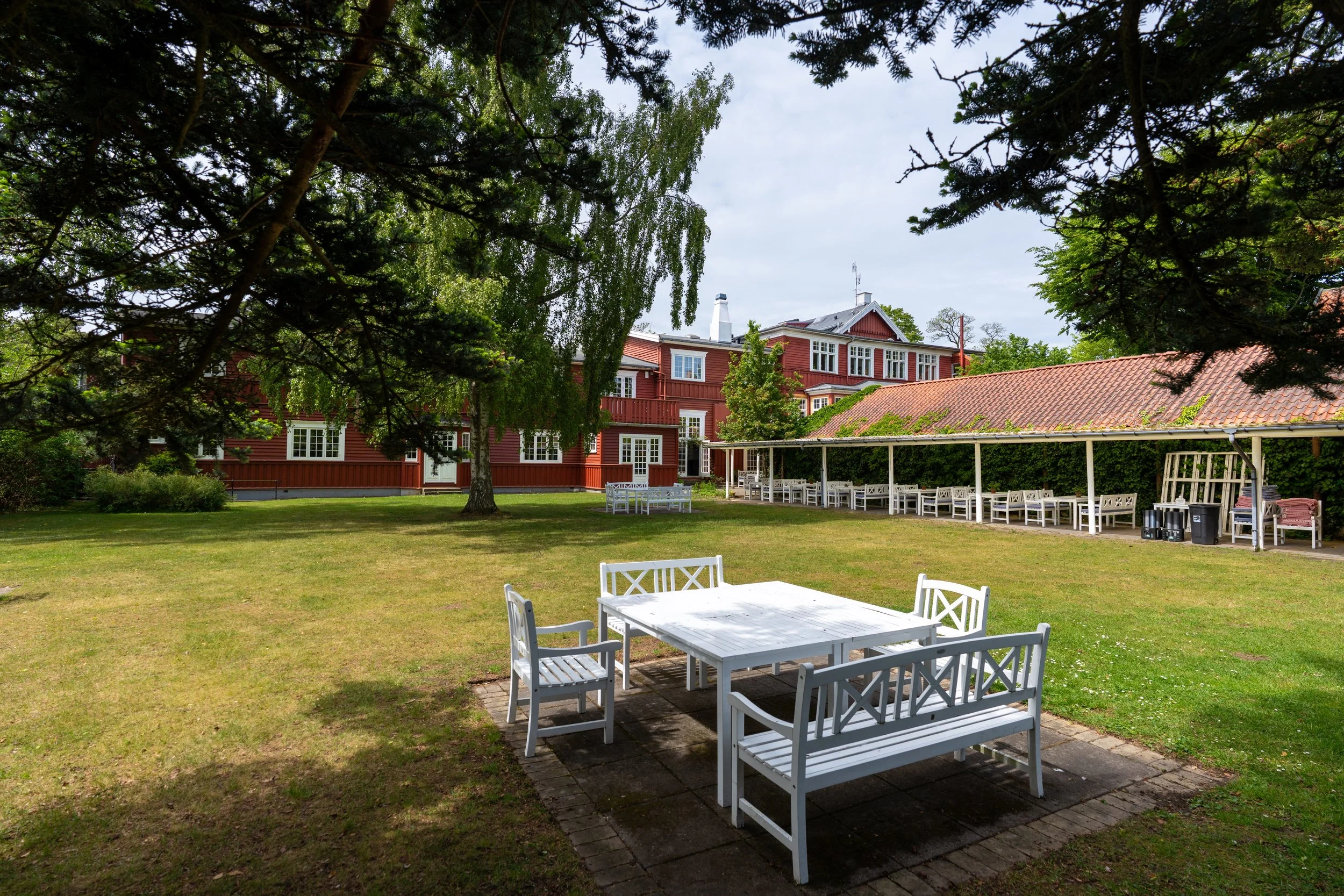 A backyard with white outdoor tables and chairs on a brick patio, surrounded by green grass and trees, with a red house and a covered patio area in the background.