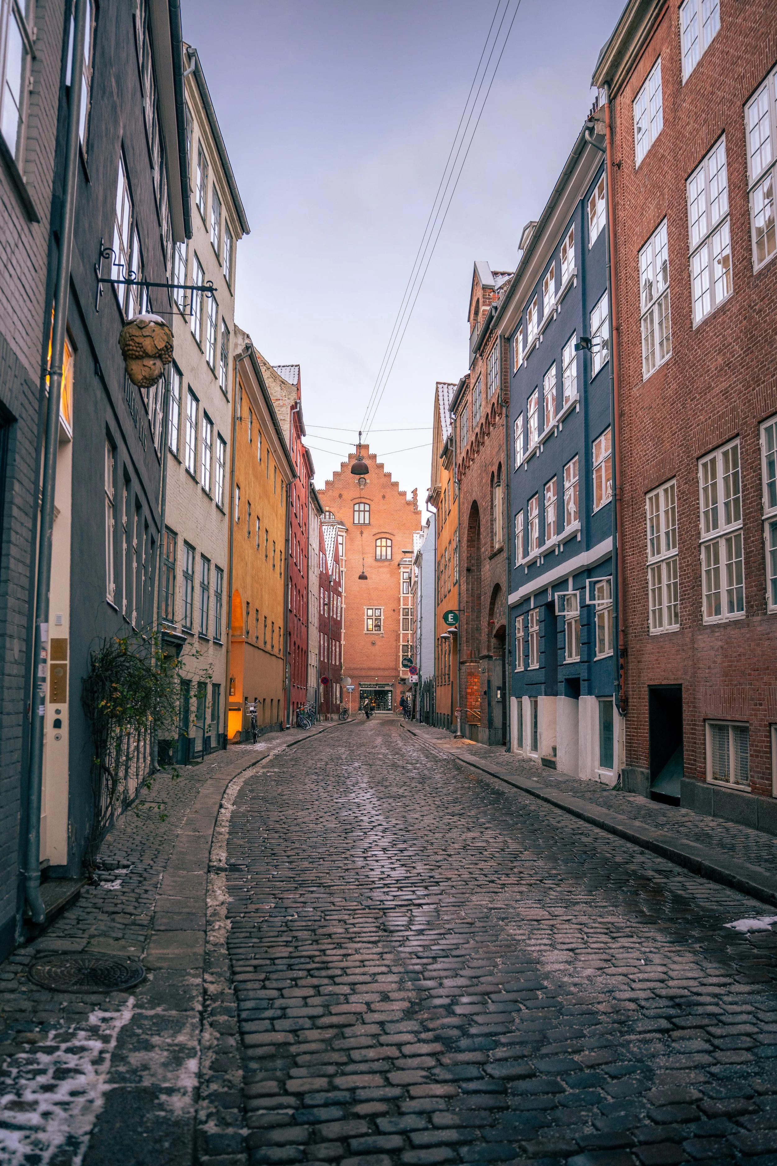 A cobblestone street lined with colorful old buildings, some with snow on the rooftops, under a clear sky in a European city.