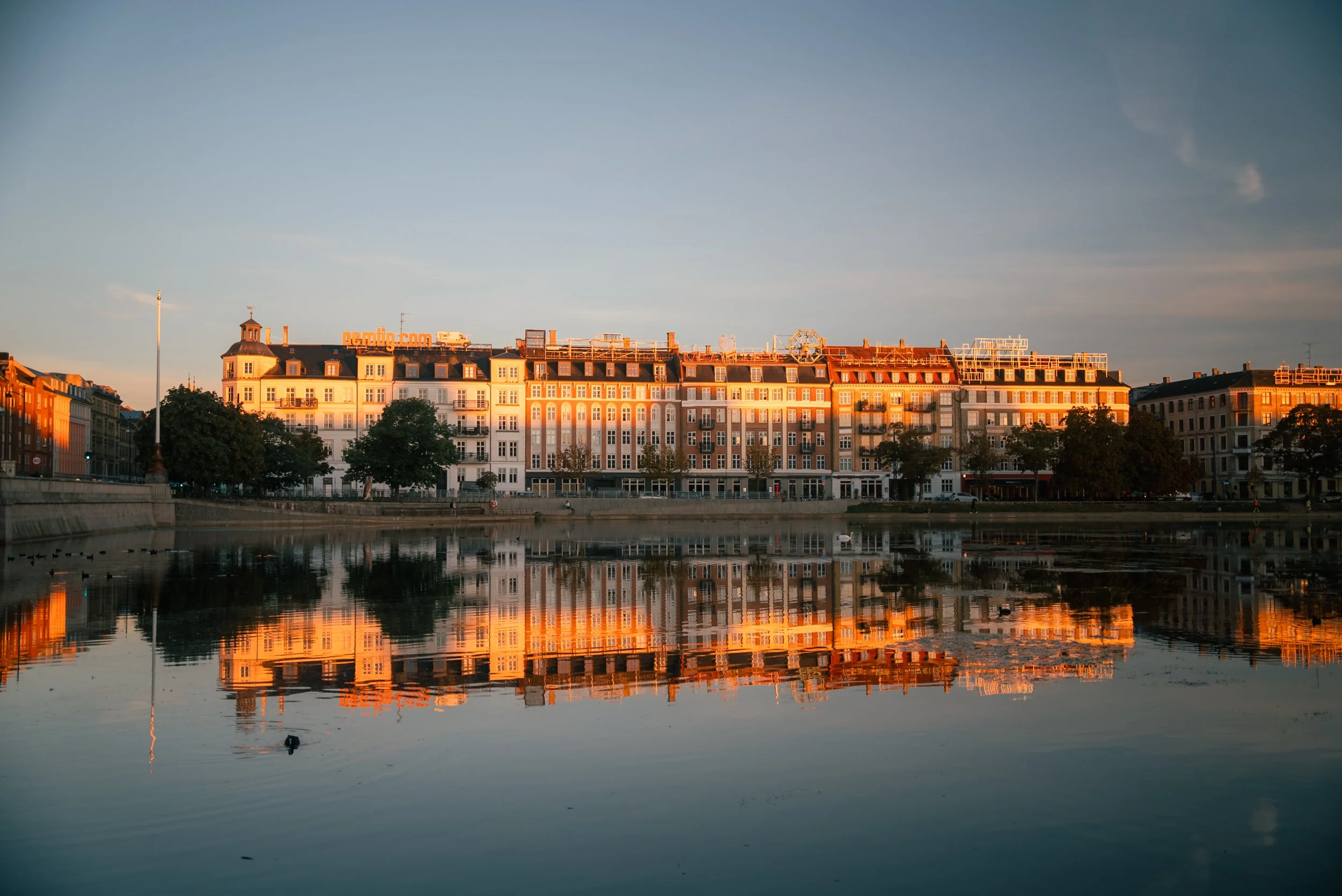 A cityscape reflecting in a river during sunset, with multistory buildings, trees, and a clear sky.