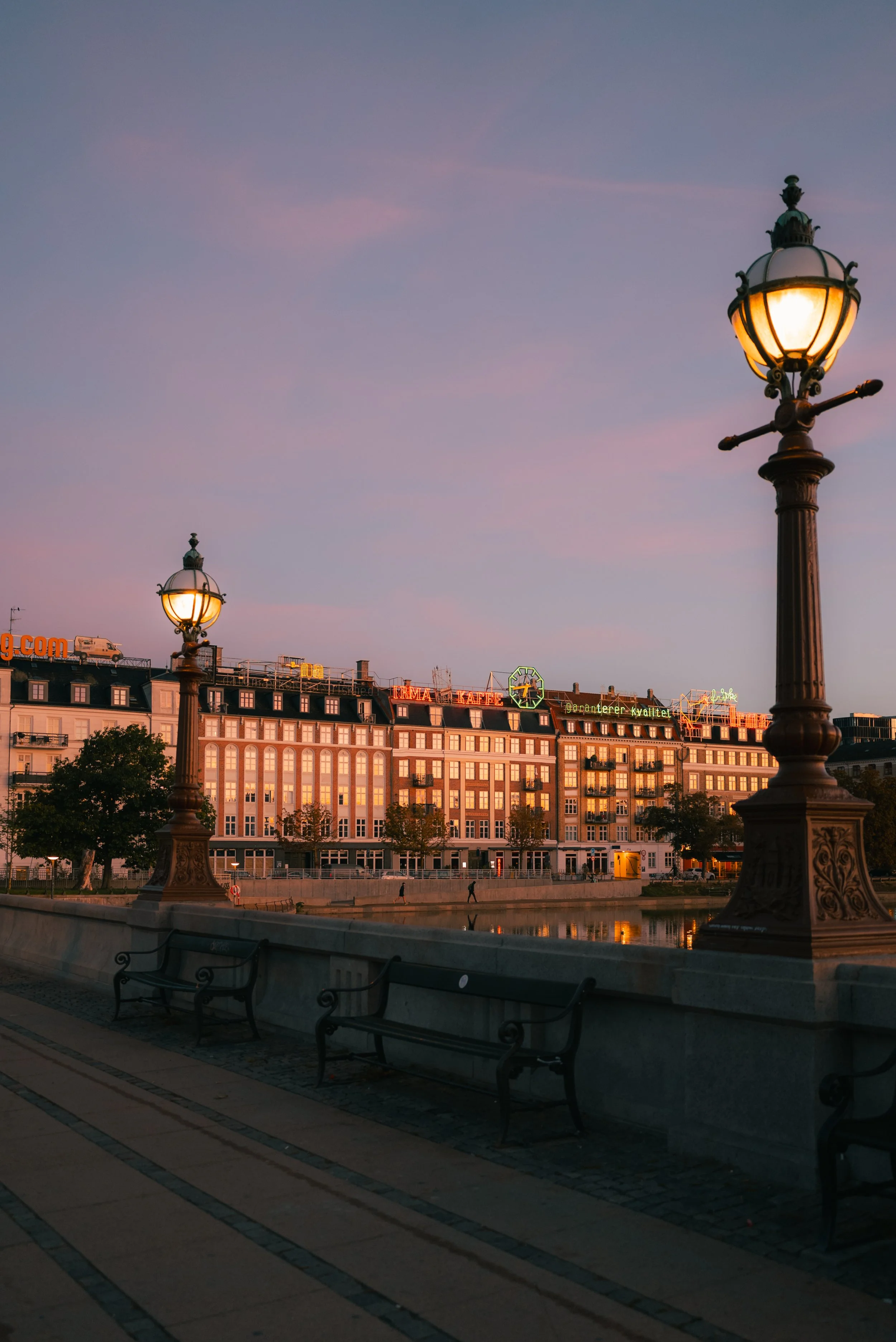A cityscape at dusk featuring ornate street lamps, a river with reflections, and buildings with neon signs, including a sign for a pharmacy and a café, in a European city.