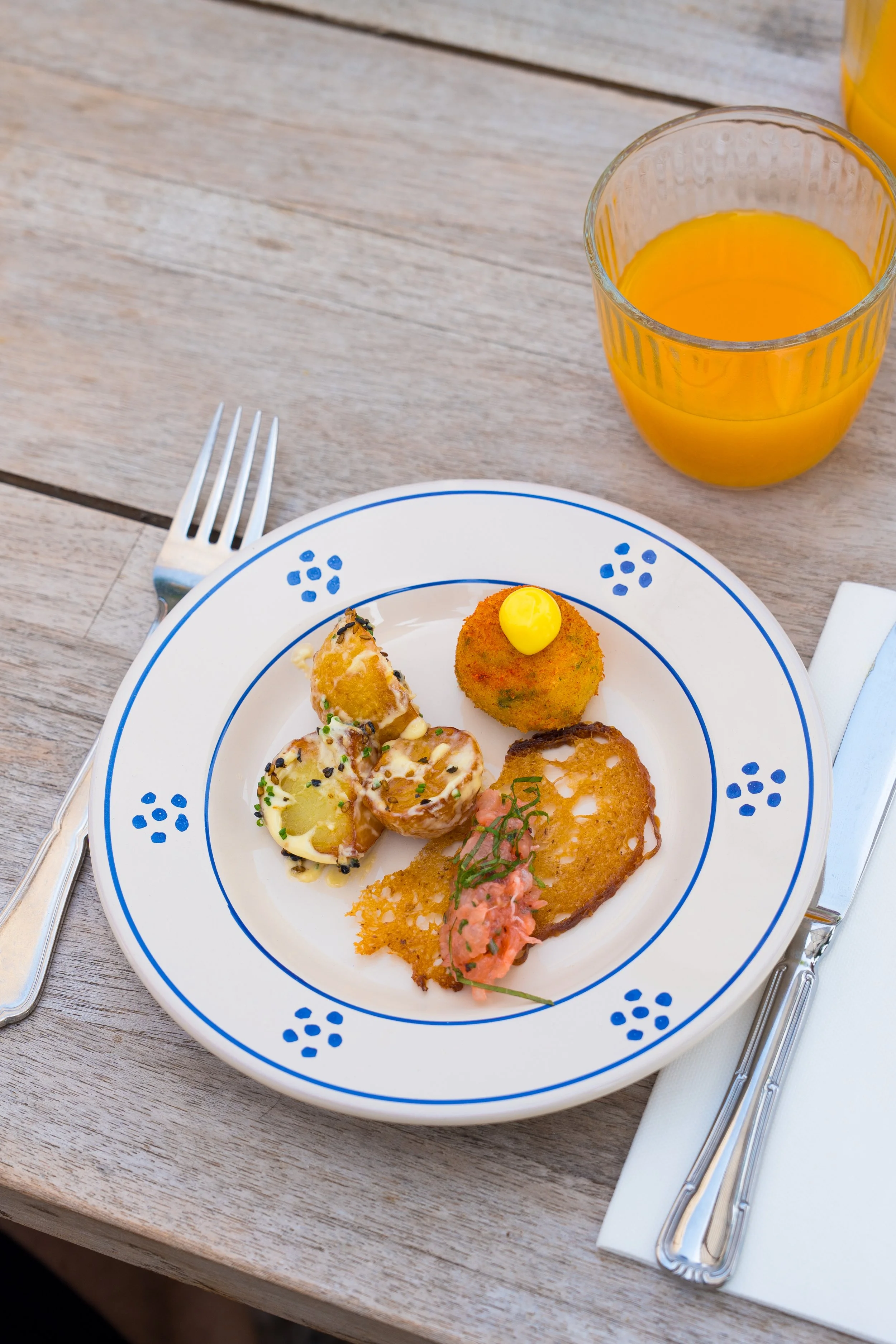 A white plate with blue decorations on a wooden table, holding various fried appetizers topped with a pink sauce and green herbs. To the right of the plate is a glass of orange juice, and a knife, fork, and white napkin are arranged next to it.