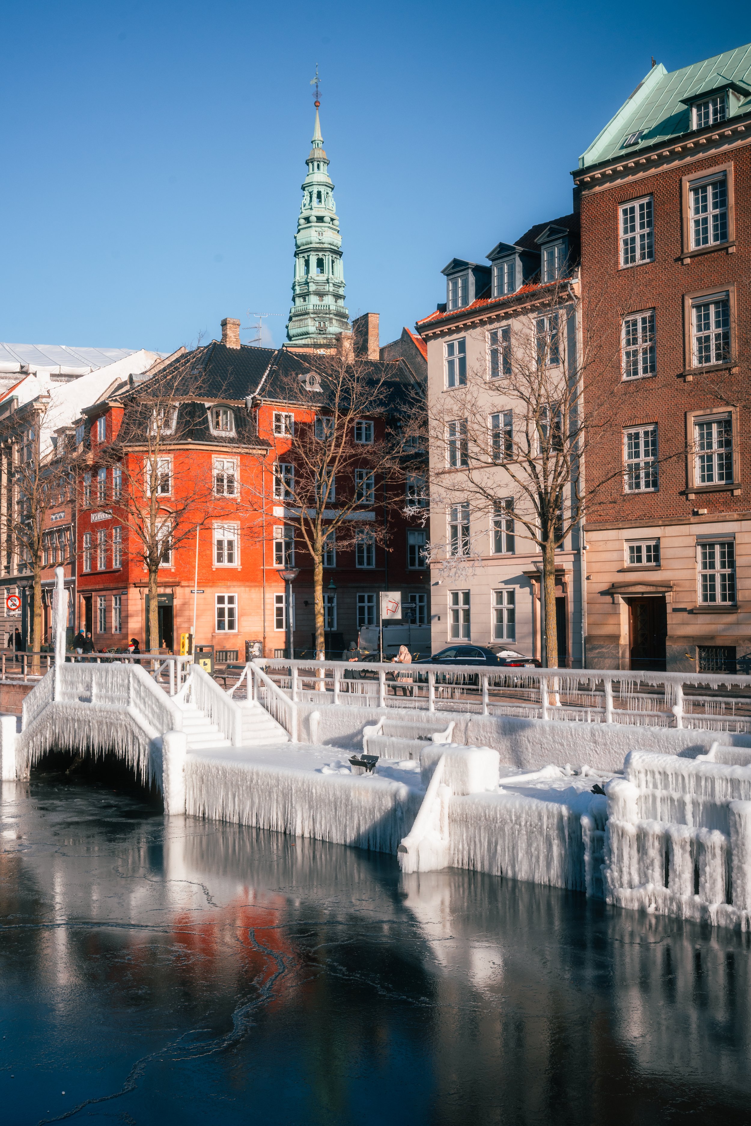 Cityscape with colorful buildings, a church steeple in the background, leafless trees, and a frozen canal with a snow-covered bridge in a winter setting on a clear sunny day.