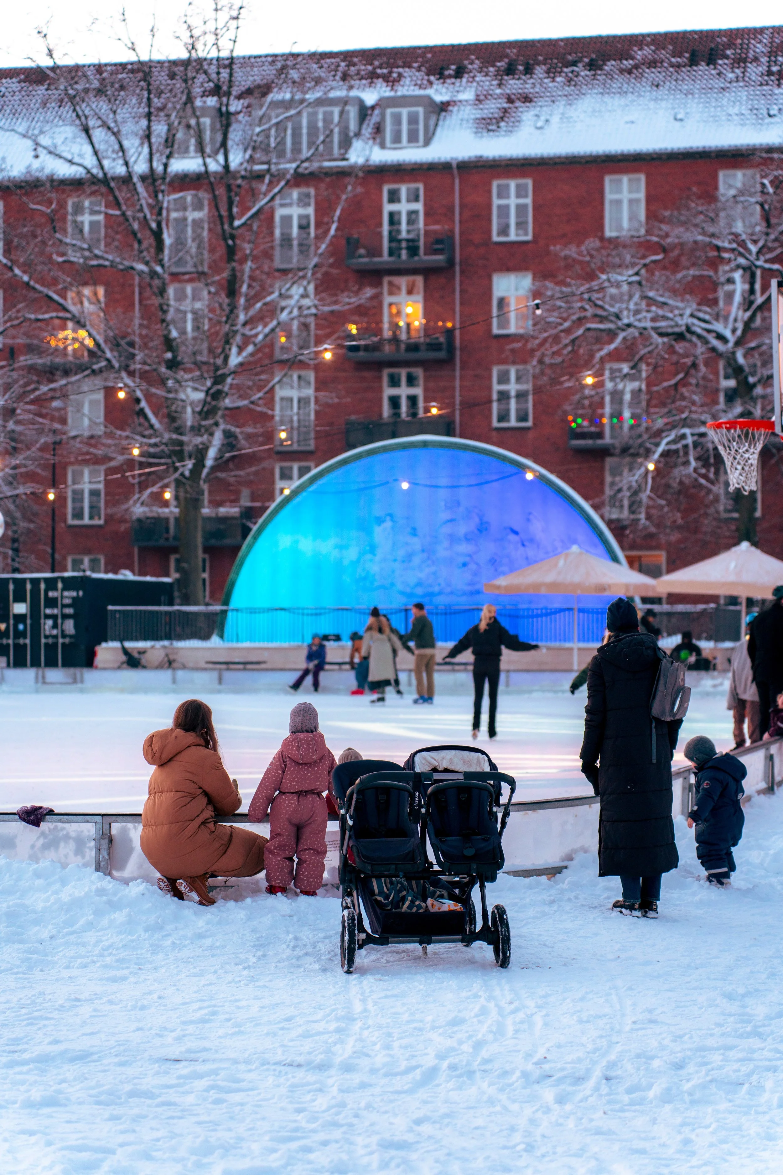 People ice skating on an outdoor rink with snow, trees, and a brick building in the background.