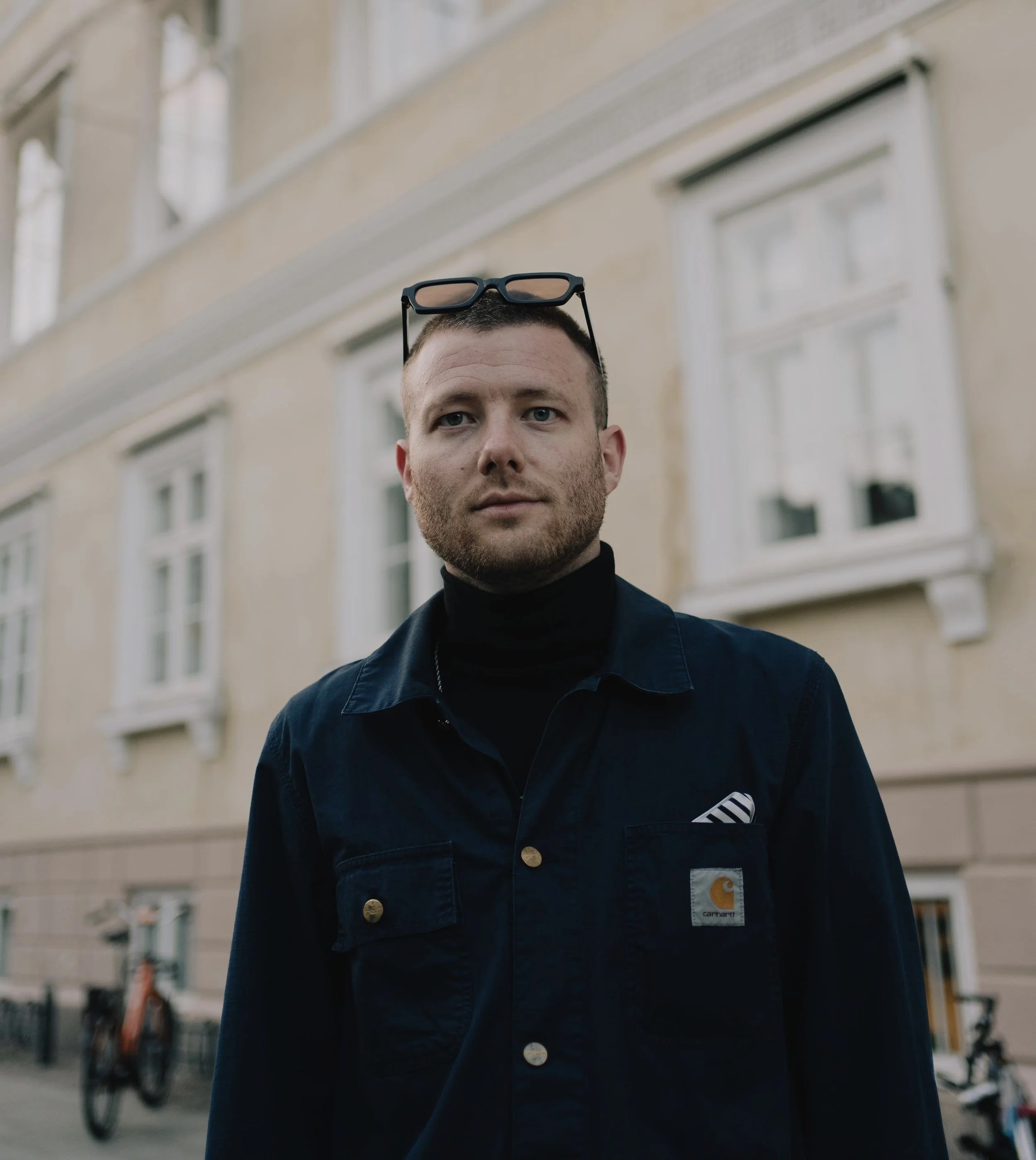 Close-up of a man's face with a dark jacket, glasses resting on his head, in an urban setting with apartment windows and bicycles in the background.