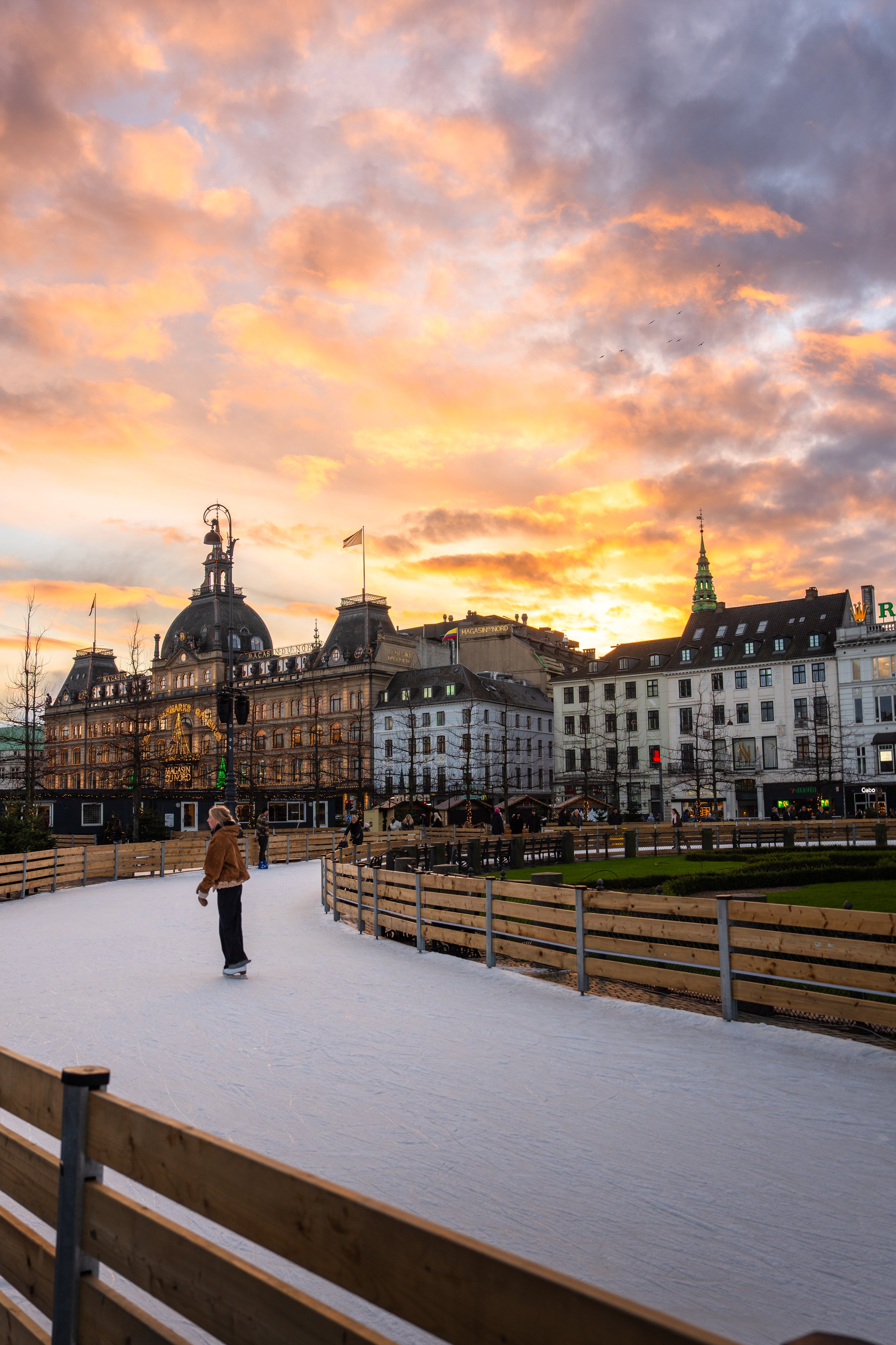 People ice skating outdoors at sunset with historic European buildings and a colorful sky in the background.