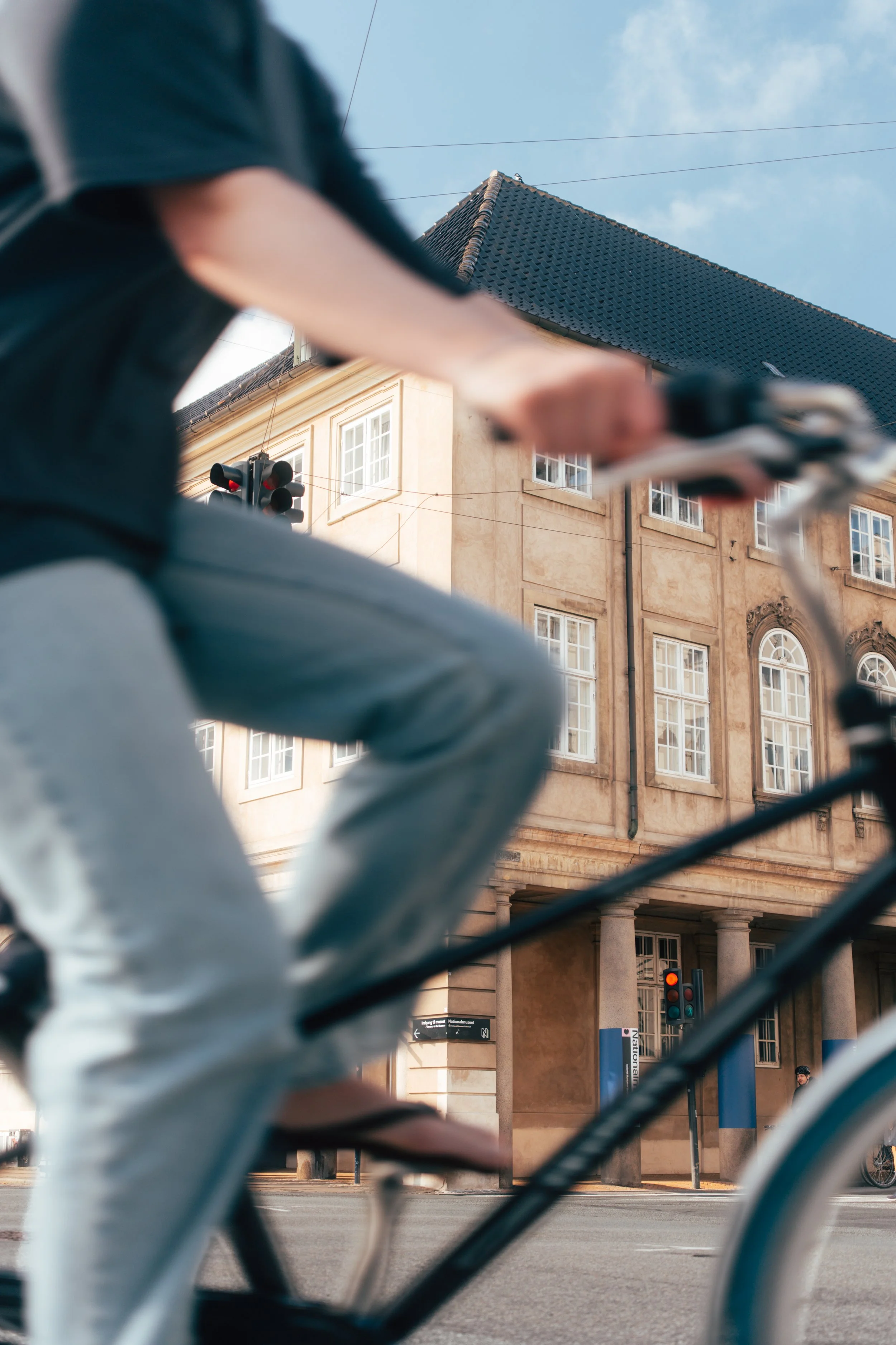 A city street scene viewed through a metal fence, with a person in a black jacket riding a bicycle in front of a historic building with large windows and a traffic light showing red, under a clear blue sky.