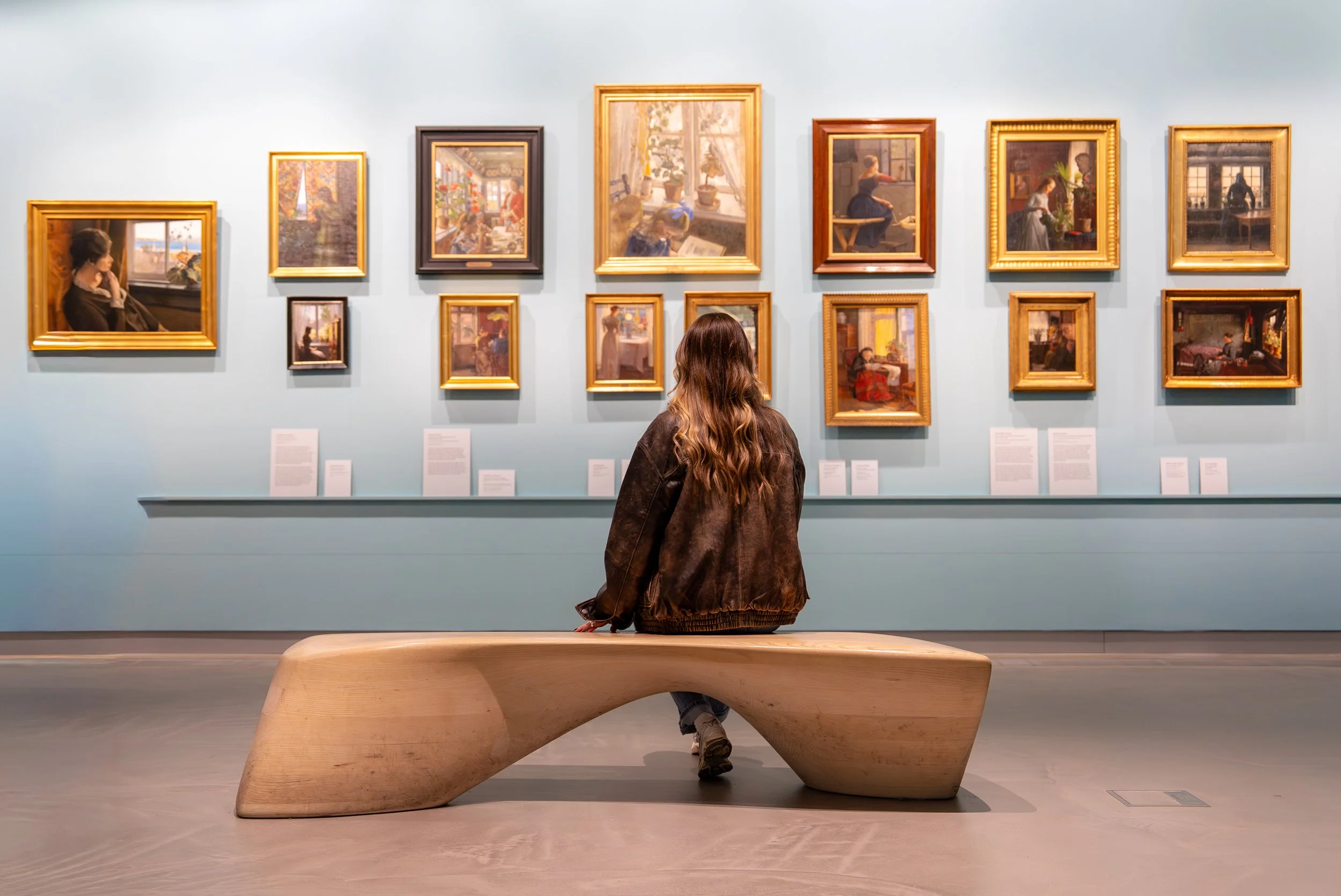 A woman with long brown hair sitting on a wooden bench in an art gallery, viewing framed paintings on a light blue wall.