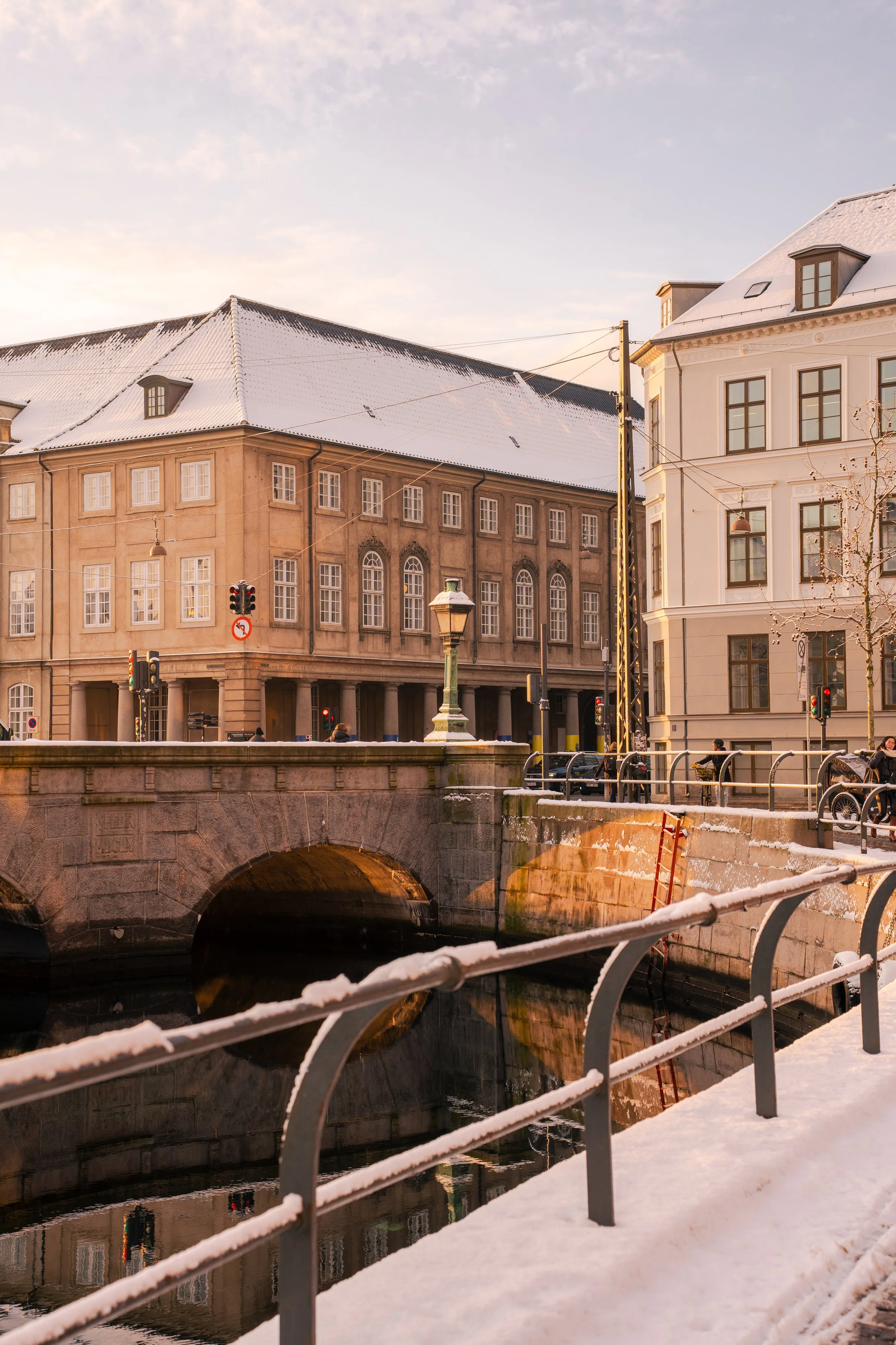 Snow-covered buildings with large windows and a bridge over a canal, winter scene with snow on the ground and rooftops, streetlights, and traffic signals.