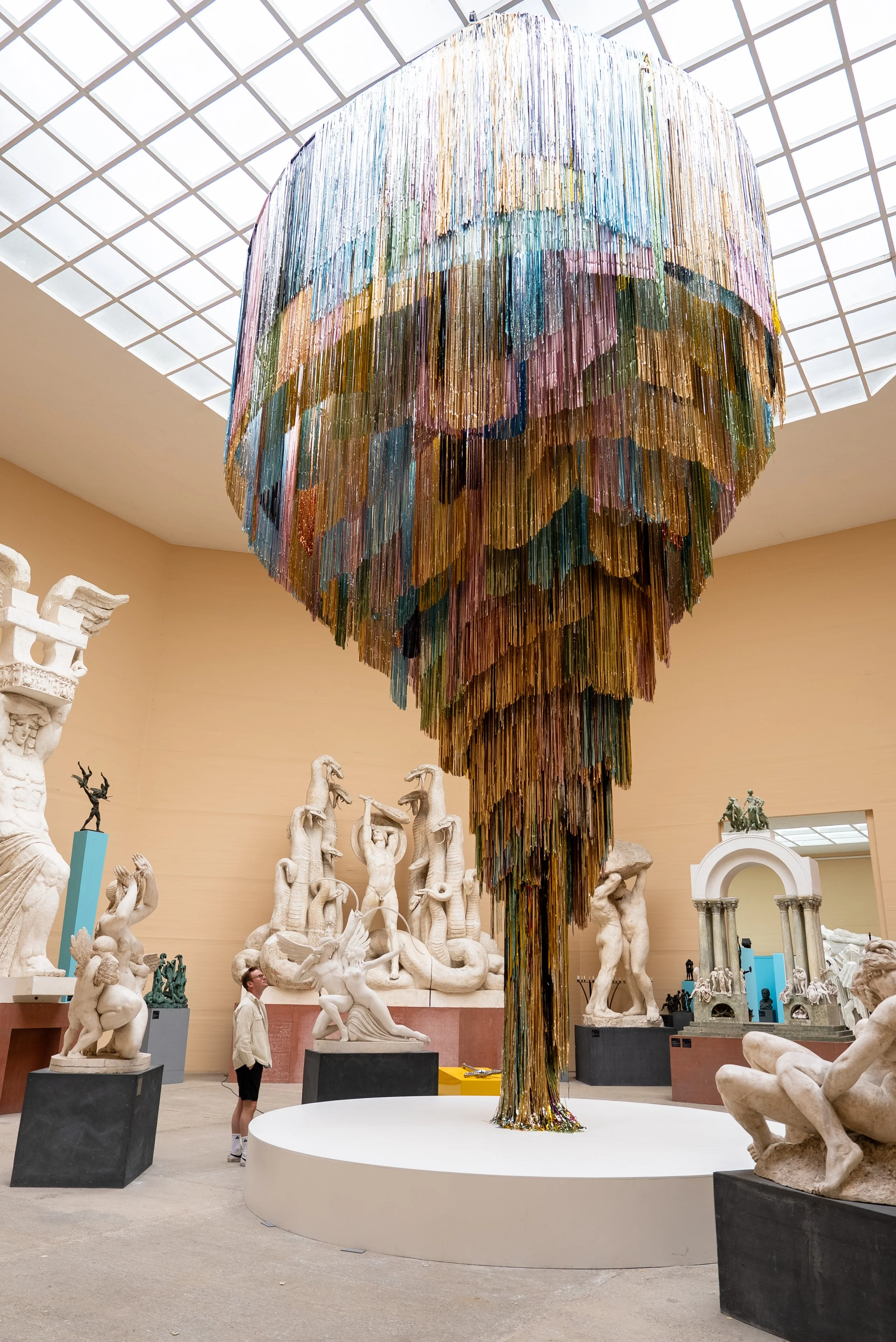 A large, colorful chandelier hangs from the ceiling of a museum gallery, surrounded by white classical sculptures on black pedestals, with a person looking at the artwork.