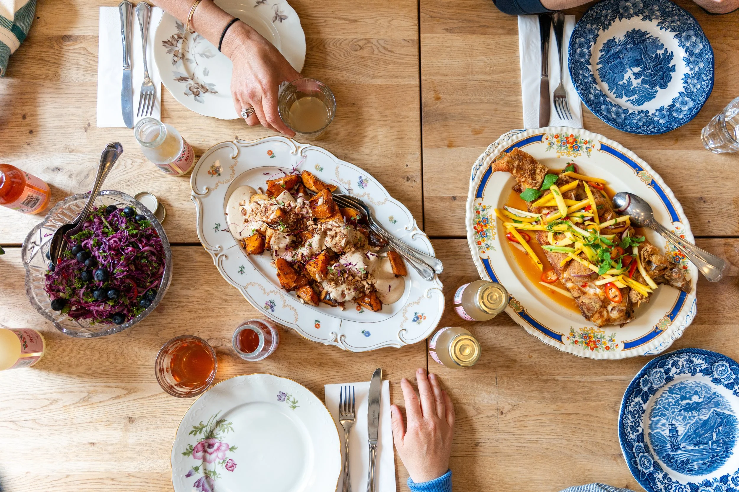 Overhead view of a wooden dining table with various dishes, drinking glasses, bottles, and cutlery. Two people are reaching for food; one has a ring and is holding a drink. The dishes include a colorful salad, roasted sweet potatoes with sauce, and a