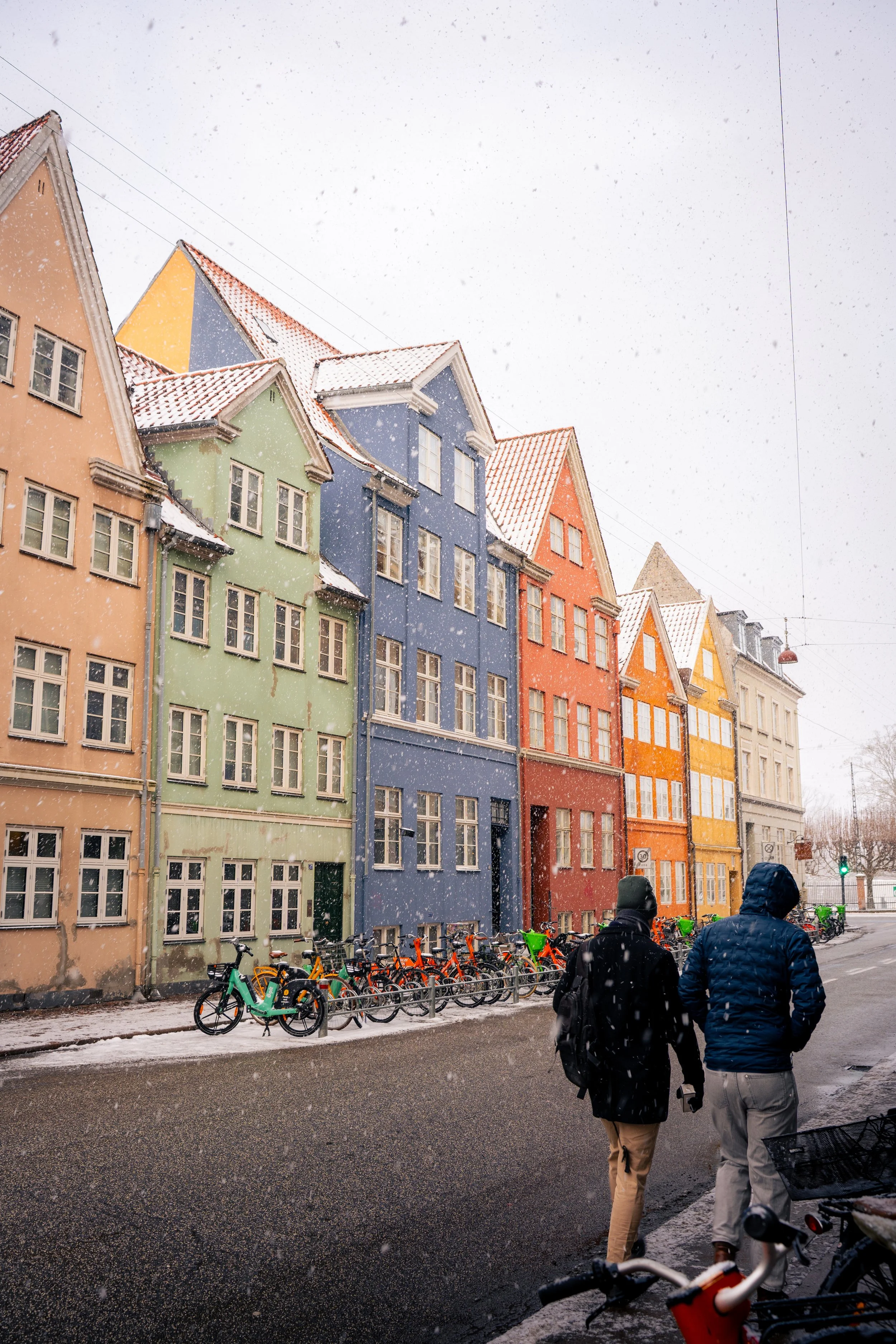 Colorful buildings along a snowy street with parked bicycles and two people walking, wearing winter jackets.