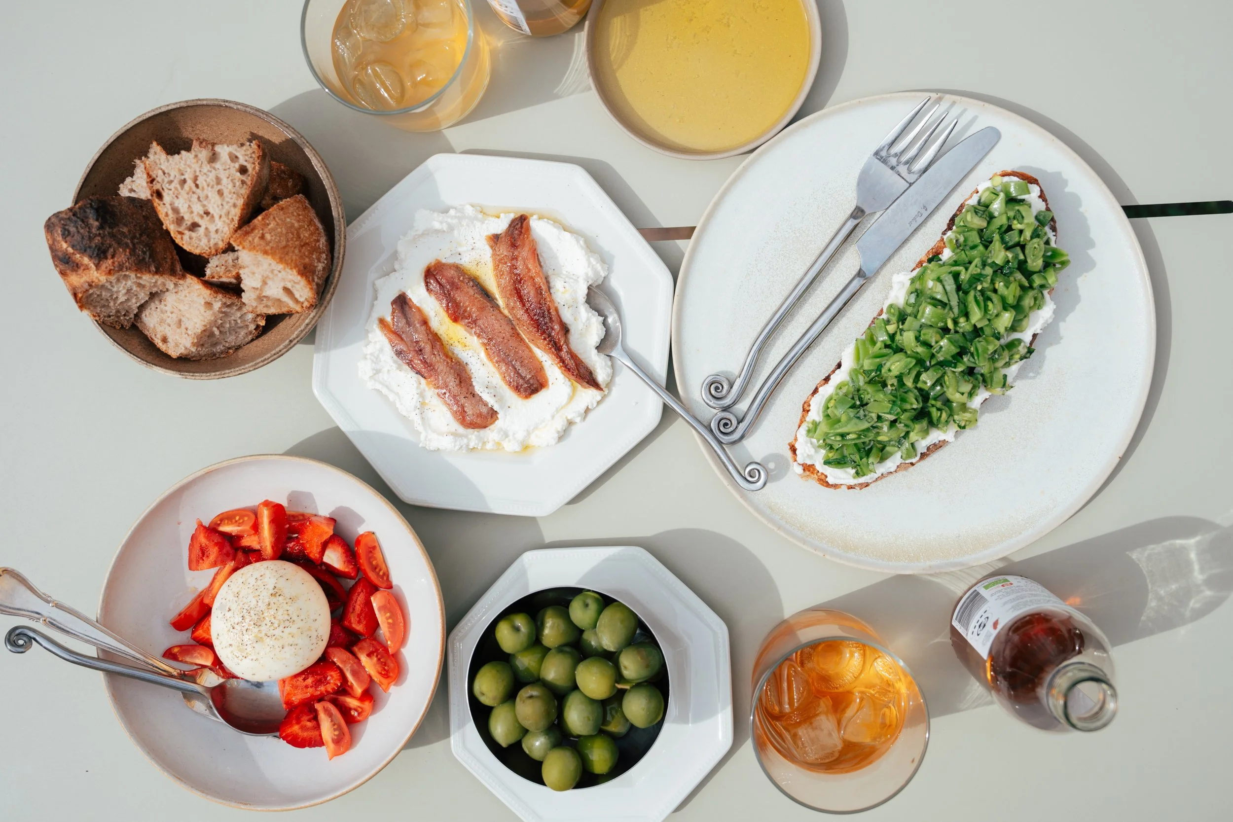 A top-down view of a breakfast spread with various dishes including toasted bread, bacon and cream cheese on bread, a poached egg with tomatoes, a green apple, a dish with chopped green vegetables, glasses of iced tea, and bowls of soup or juice.