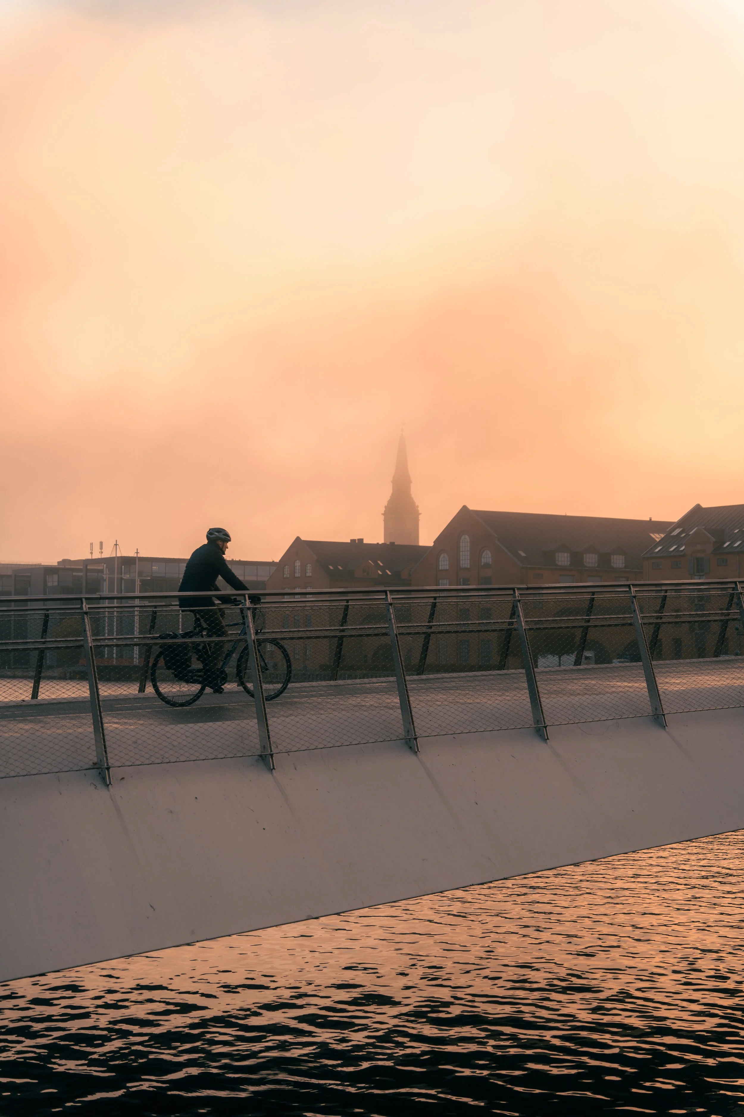 A person riding a bicycle on a bridge at sunset with a cityscape and church tower in the background.