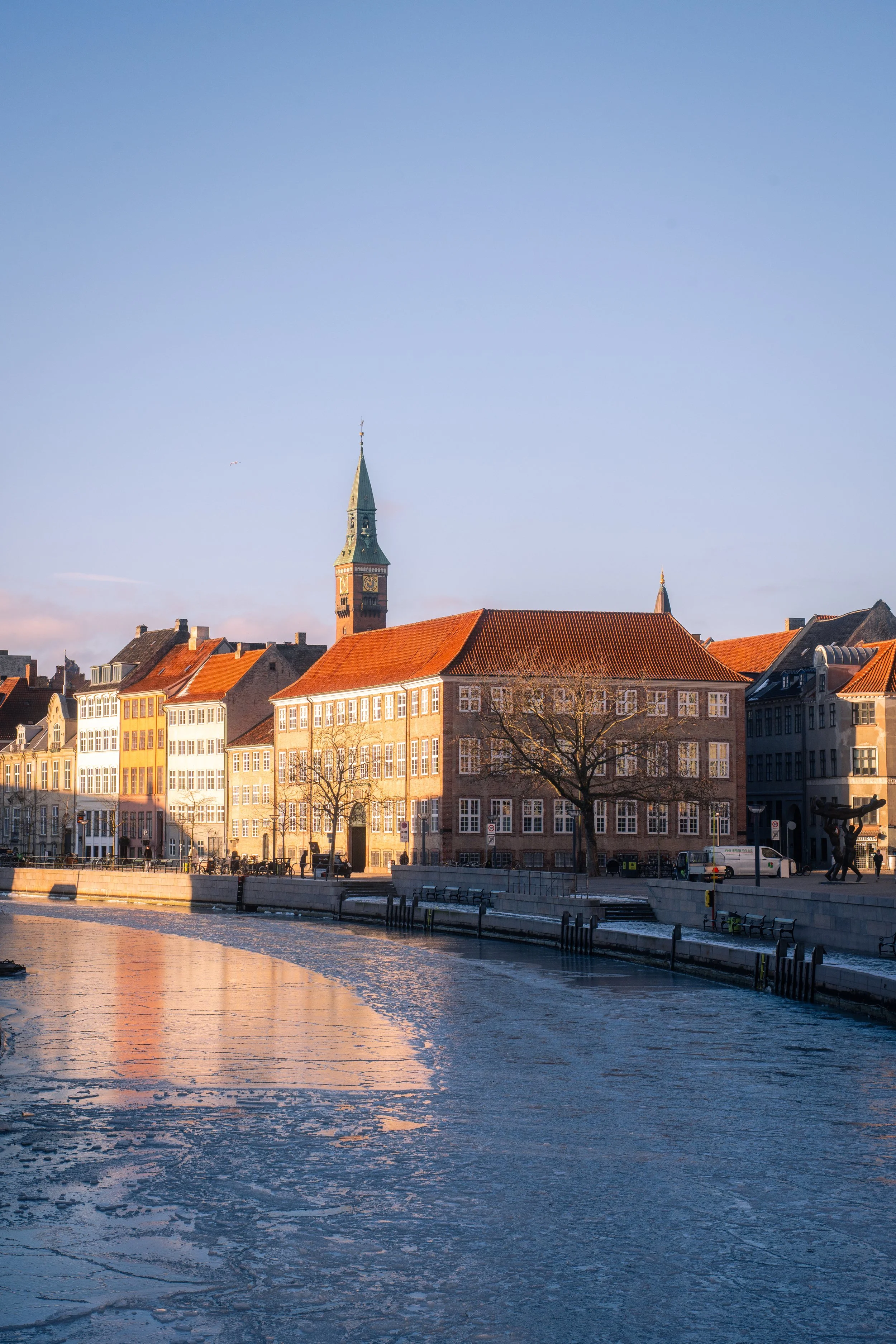 View of historic European buildings along a riverbank with a church spire in the background under a clear blue sky.