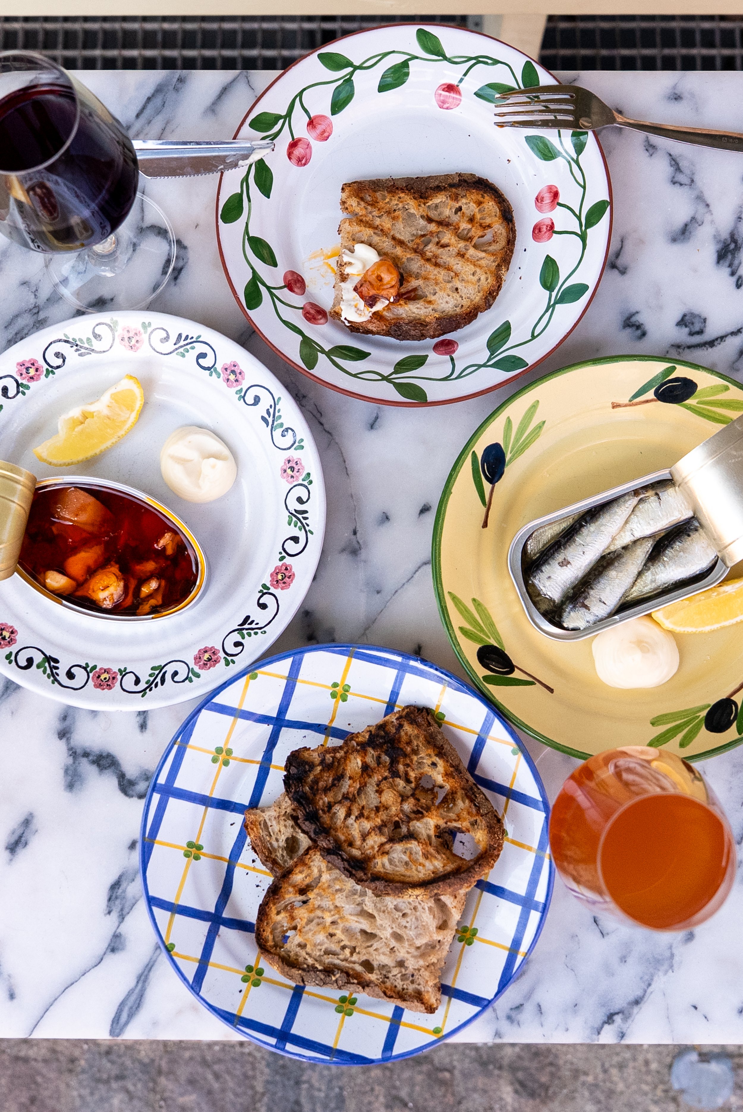 A marble table with various plates of food, including slices of bread, a bowl of mussels in tomato sauce, a lemon wedge, and a glass of red wine.
