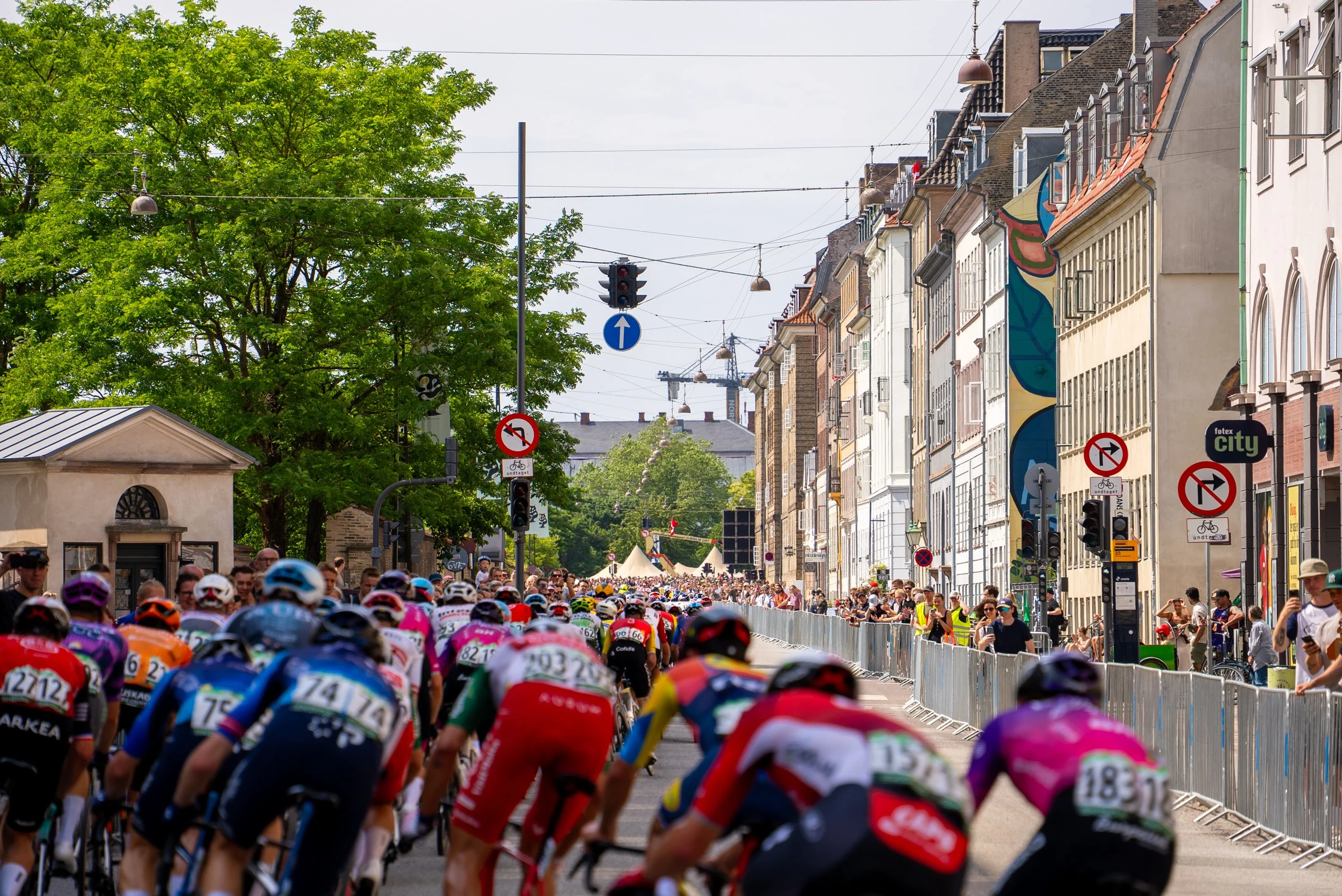 A large crowd of cyclists participating in a race on a city street lined with colorful buildings and trees.