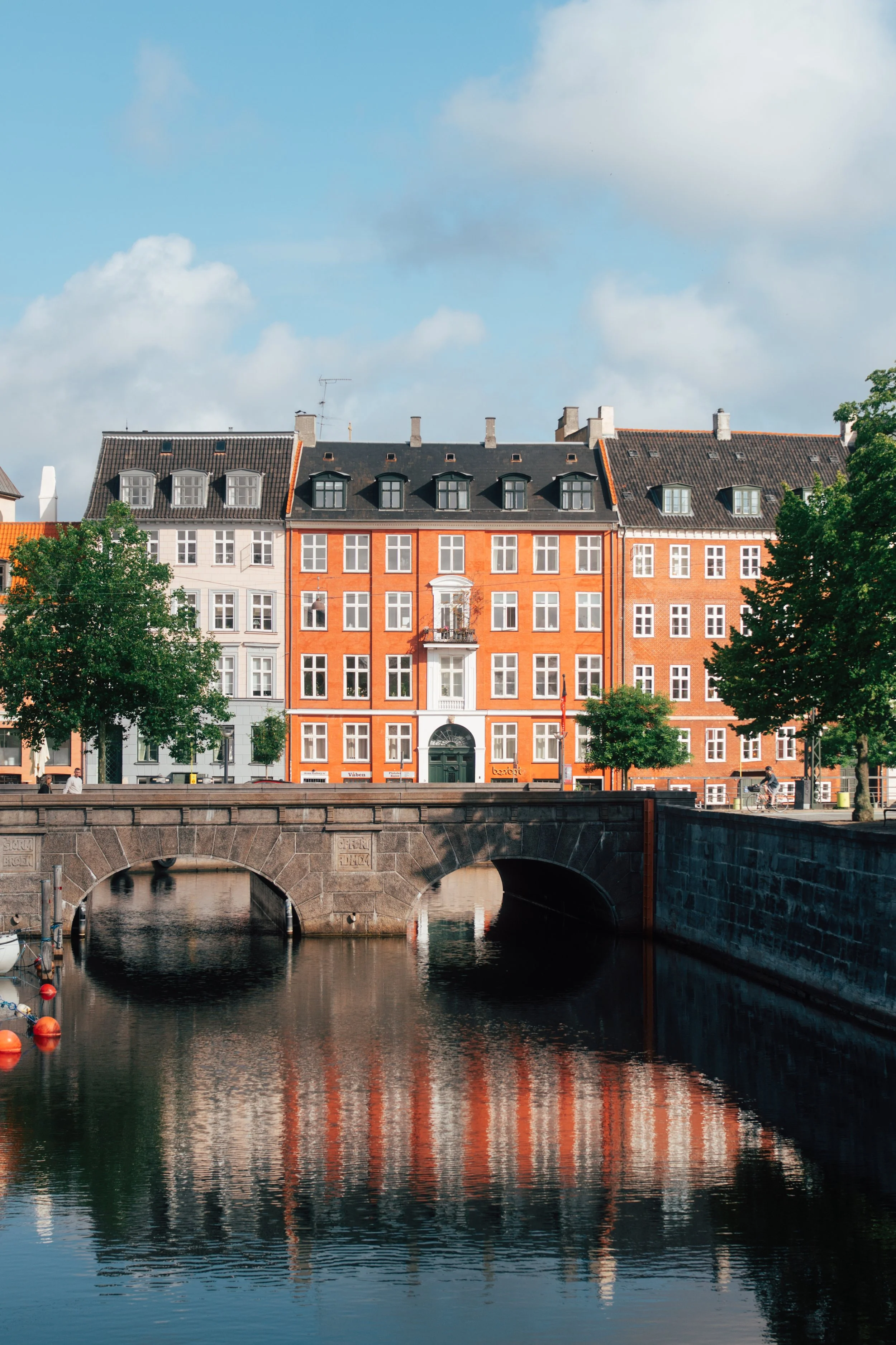 A waterfront scene in a European city featuring historic orange and beige buildings with dark roofs, a bridge over a canal, and boats moored along the water, under a partly cloudy sky.