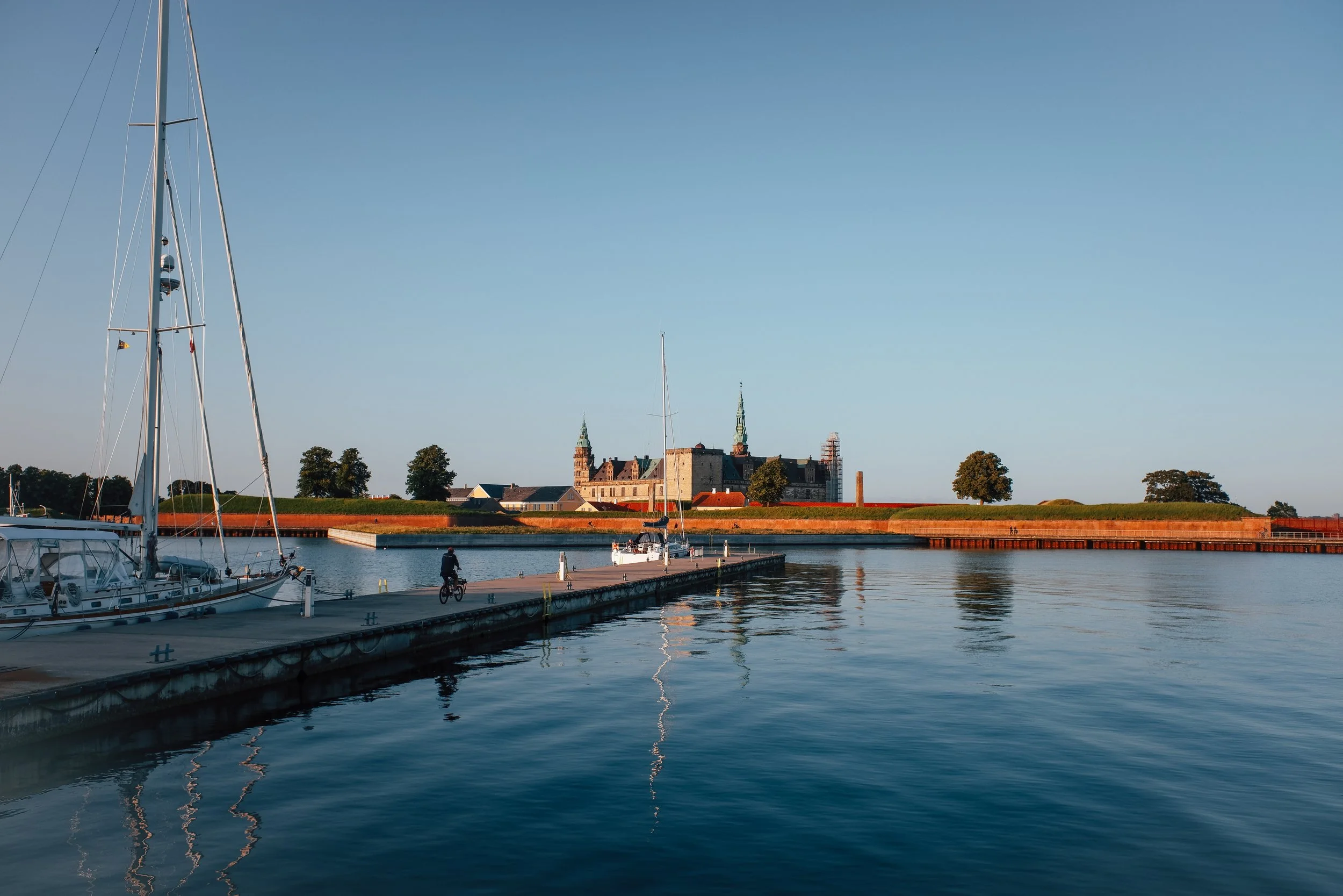 Boats docked at a marina with a historic building and trees in the background under a clear blue sky.