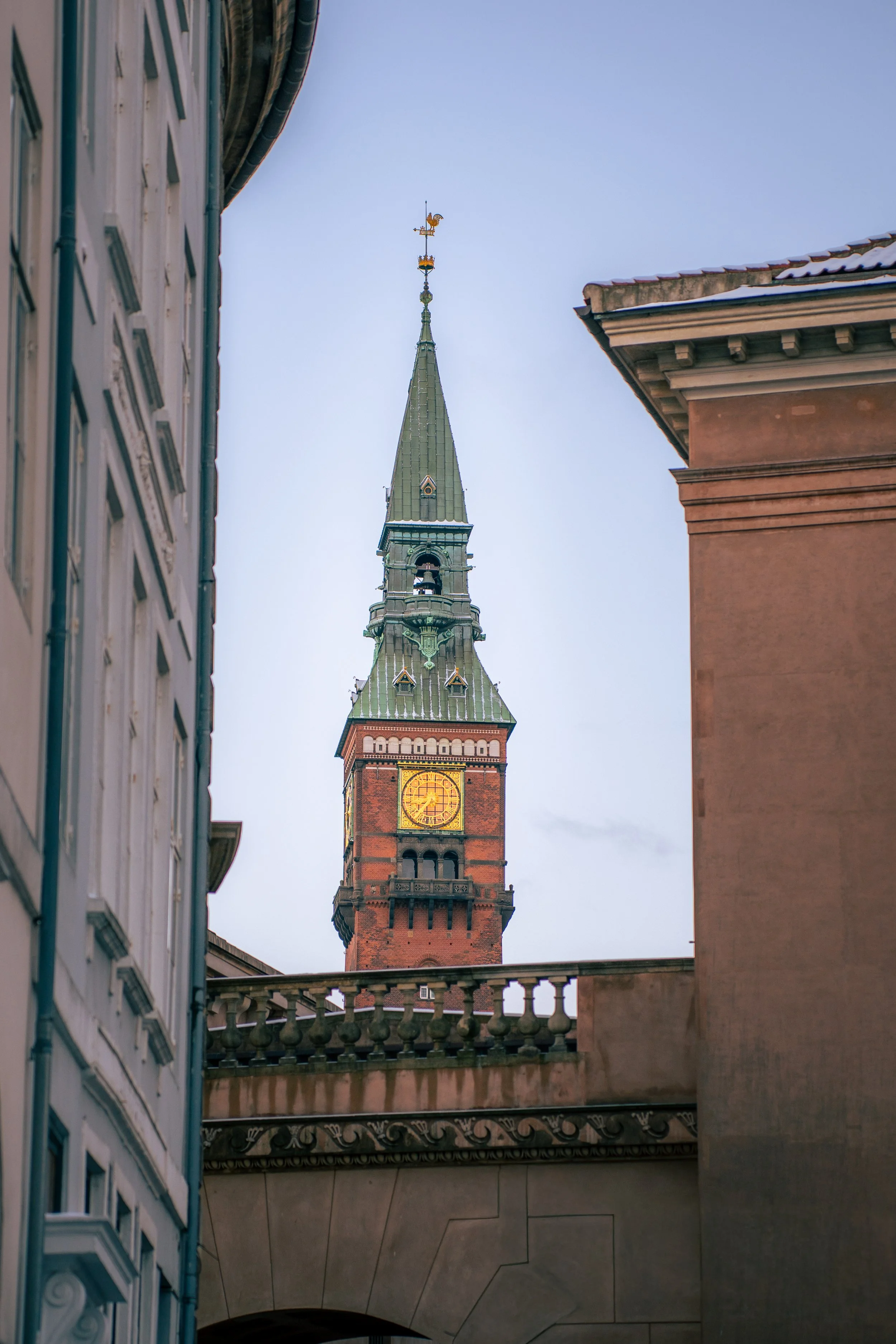 The image shows a tall clock tower with a green spire, flanked by two buildings on either side, and a clear blue sky in the background.