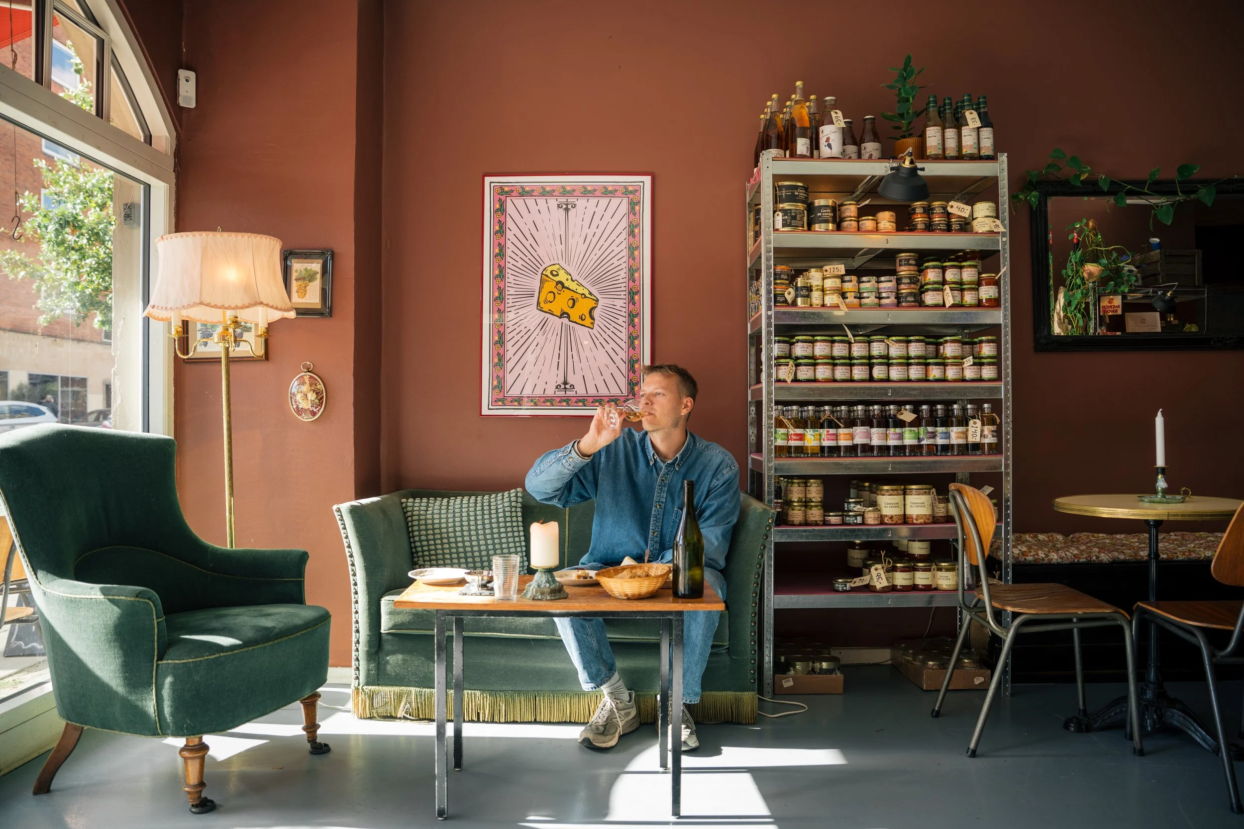 A man is sitting at a table inside a cozy cafe, drinking a glass of wine. The cafe has vintage-style green chairs, a tall standing lamp, and a shelf filled with jars and bottles. There is a picture of cheese on the wall, and sunlight streams in throu