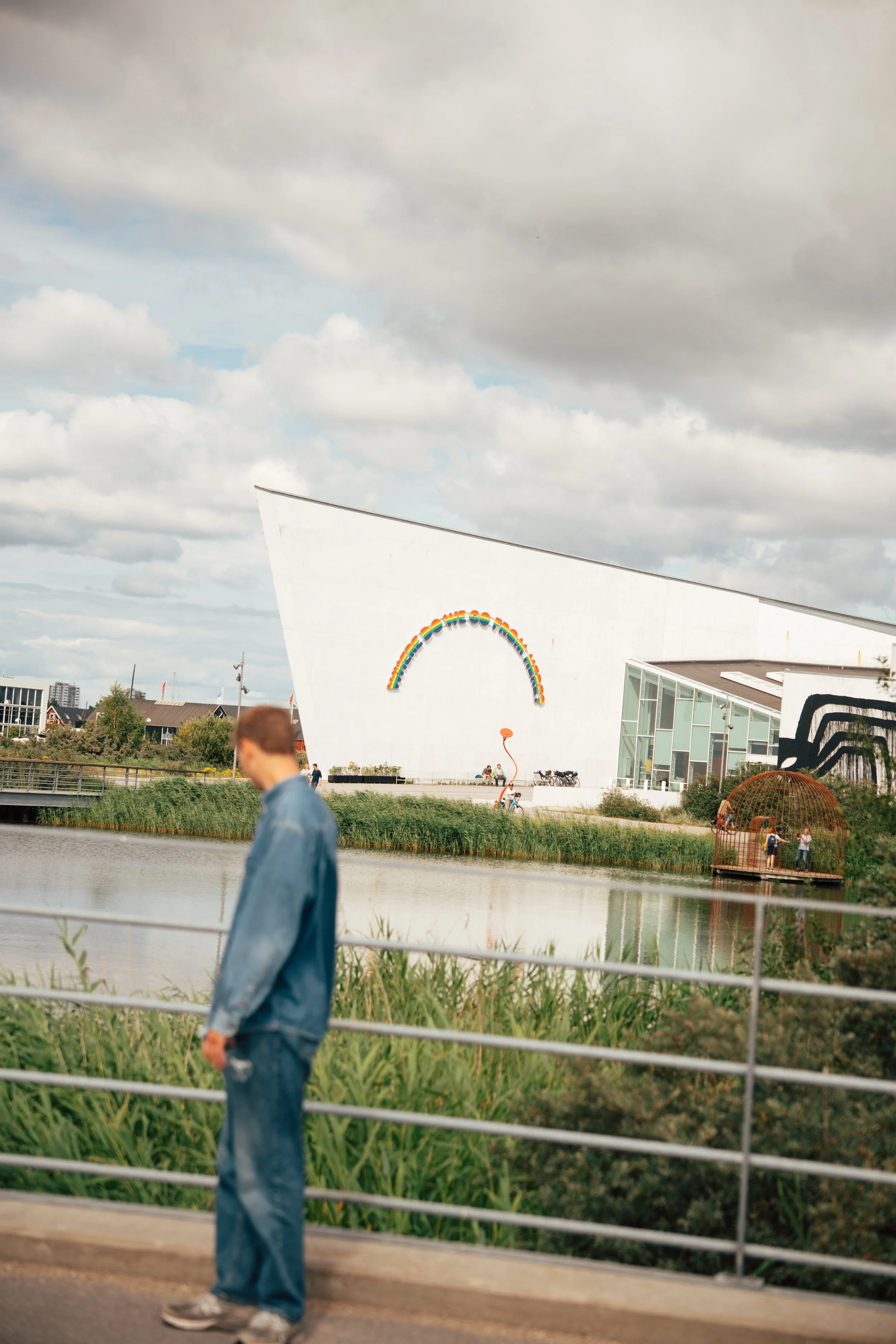 A person in a denim jacket walking along a path by a body of water with a modern white building with a rainbow design on it in the background, under cloudy skies.