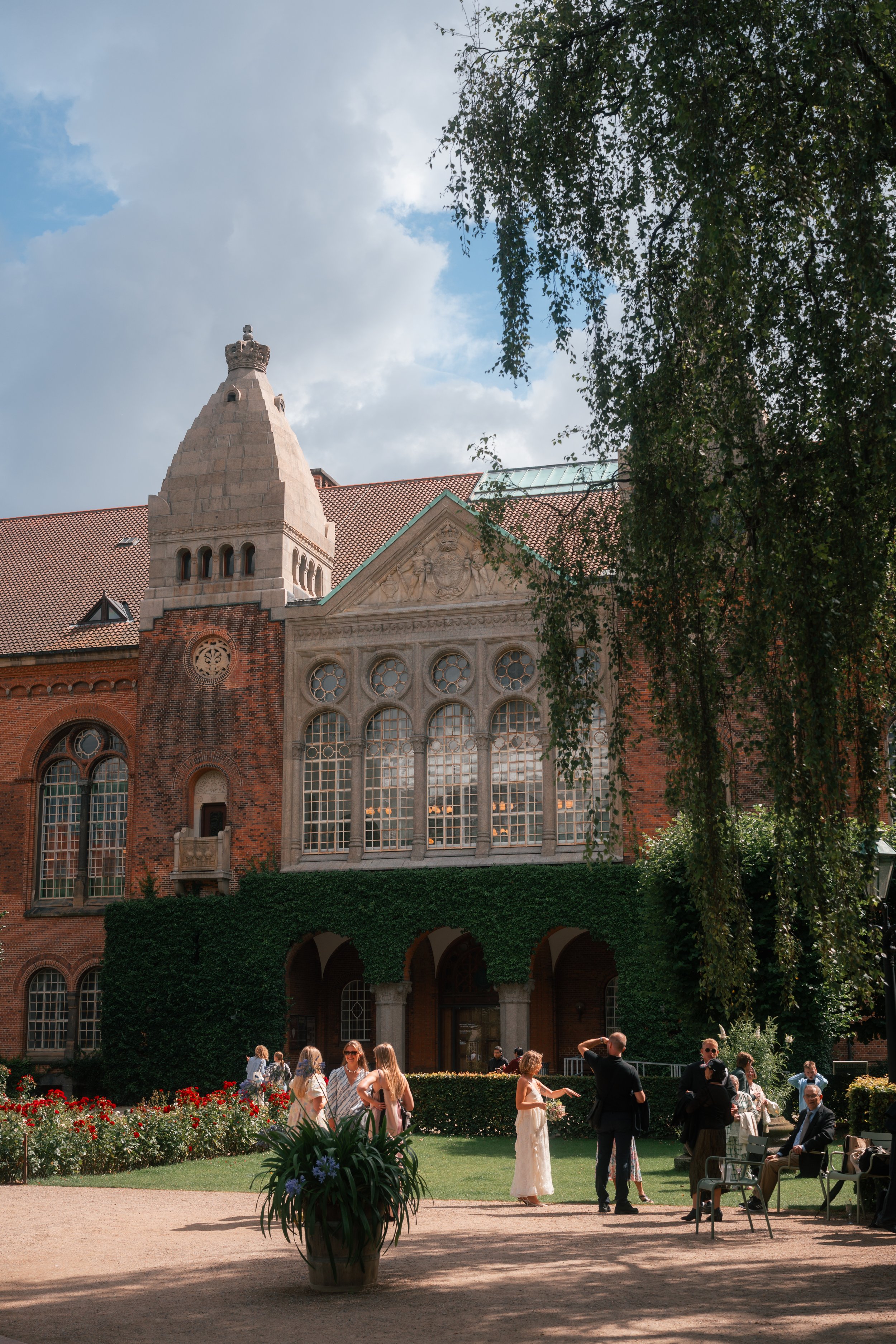 A group of people gathered outside a historic building with red brick and stone architecture, surrounded by a garden with flowers and greenery, under a partly cloudy sky.