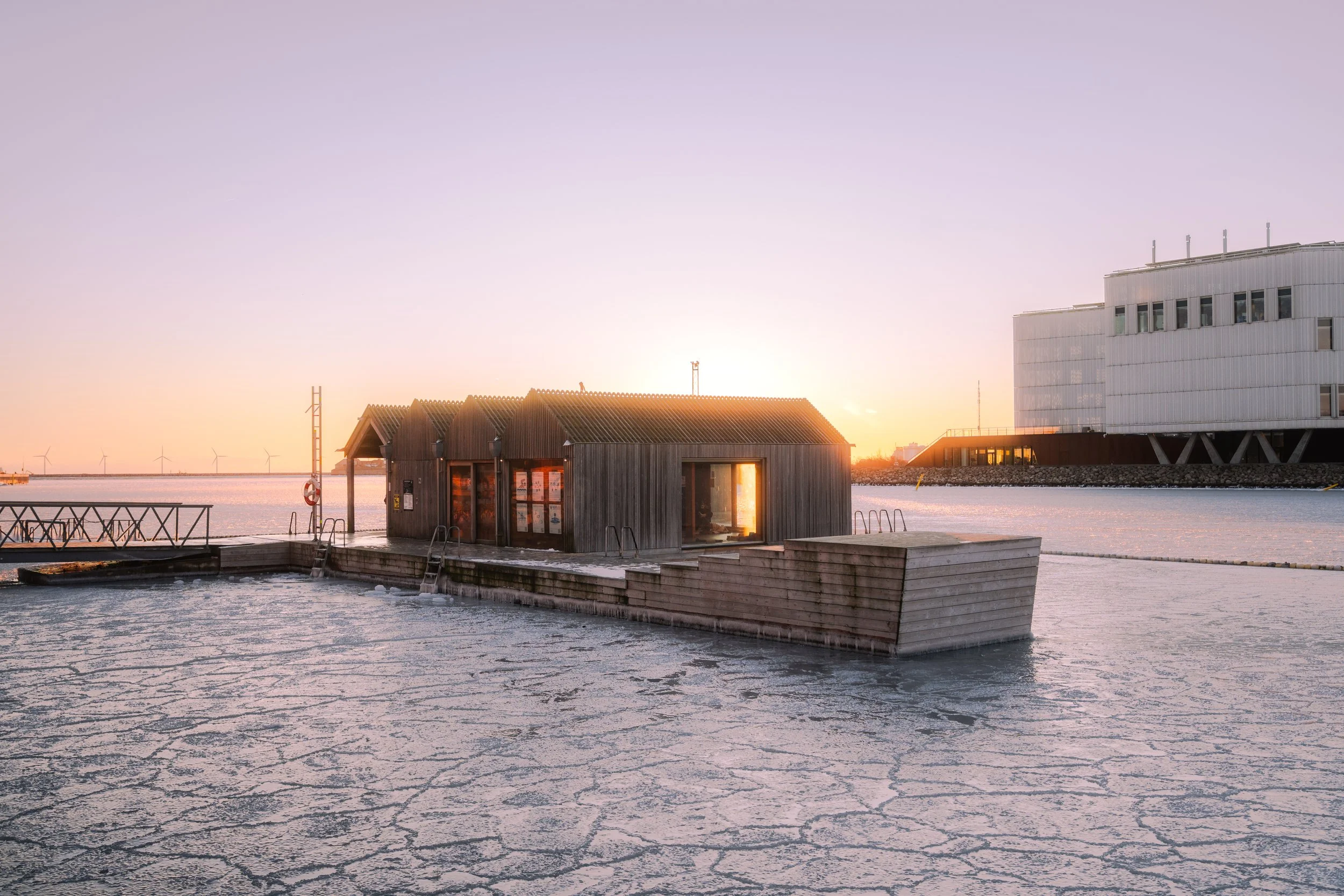 Sunset over icy pier with a small wooden building, modern building in the background, and wind turbines in the distance.