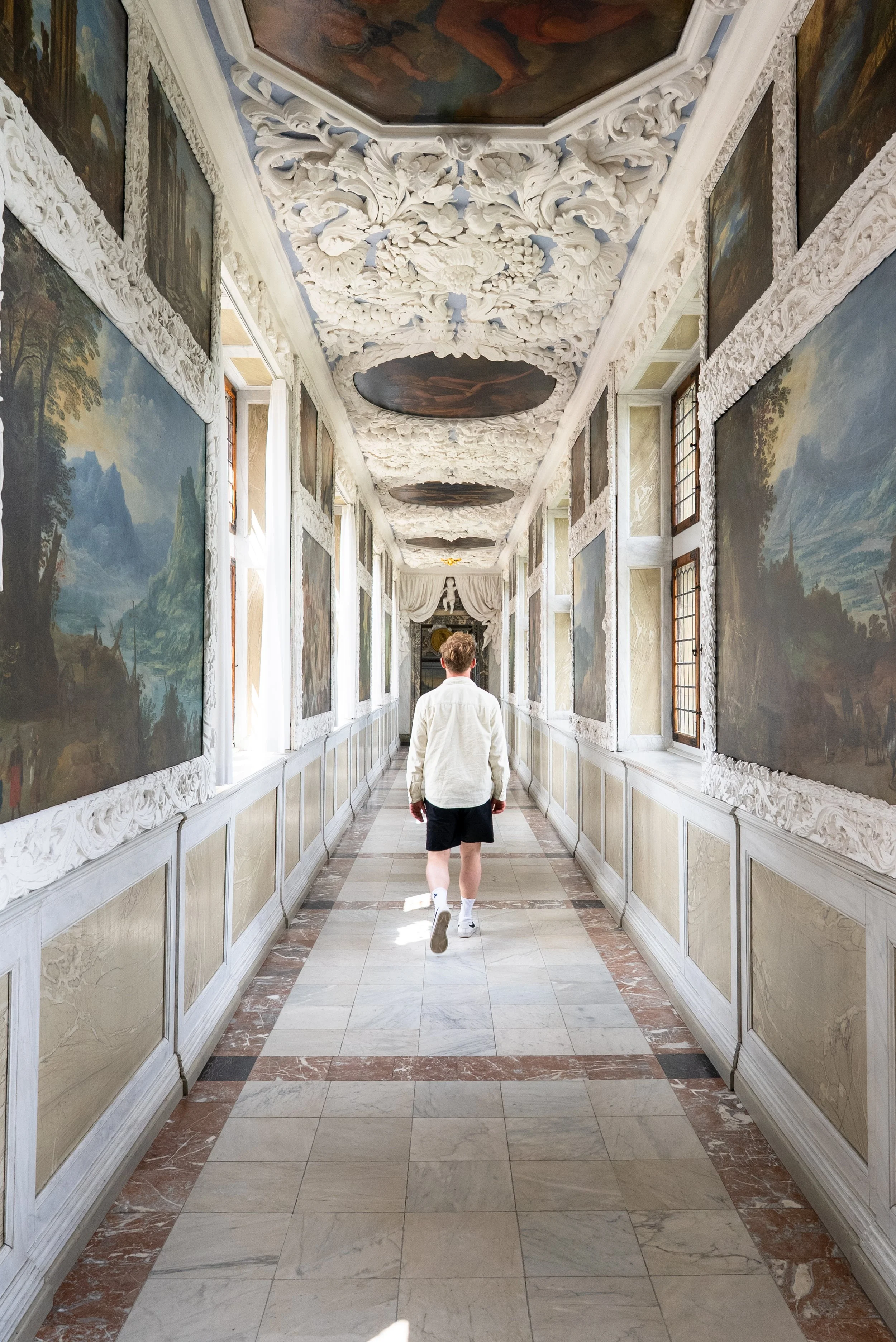 A person walking down a decorated hallway with ornate white carved walls, landscape paintings, large windows, and a ceiling with intricate white floral patterns and oval painted sections.