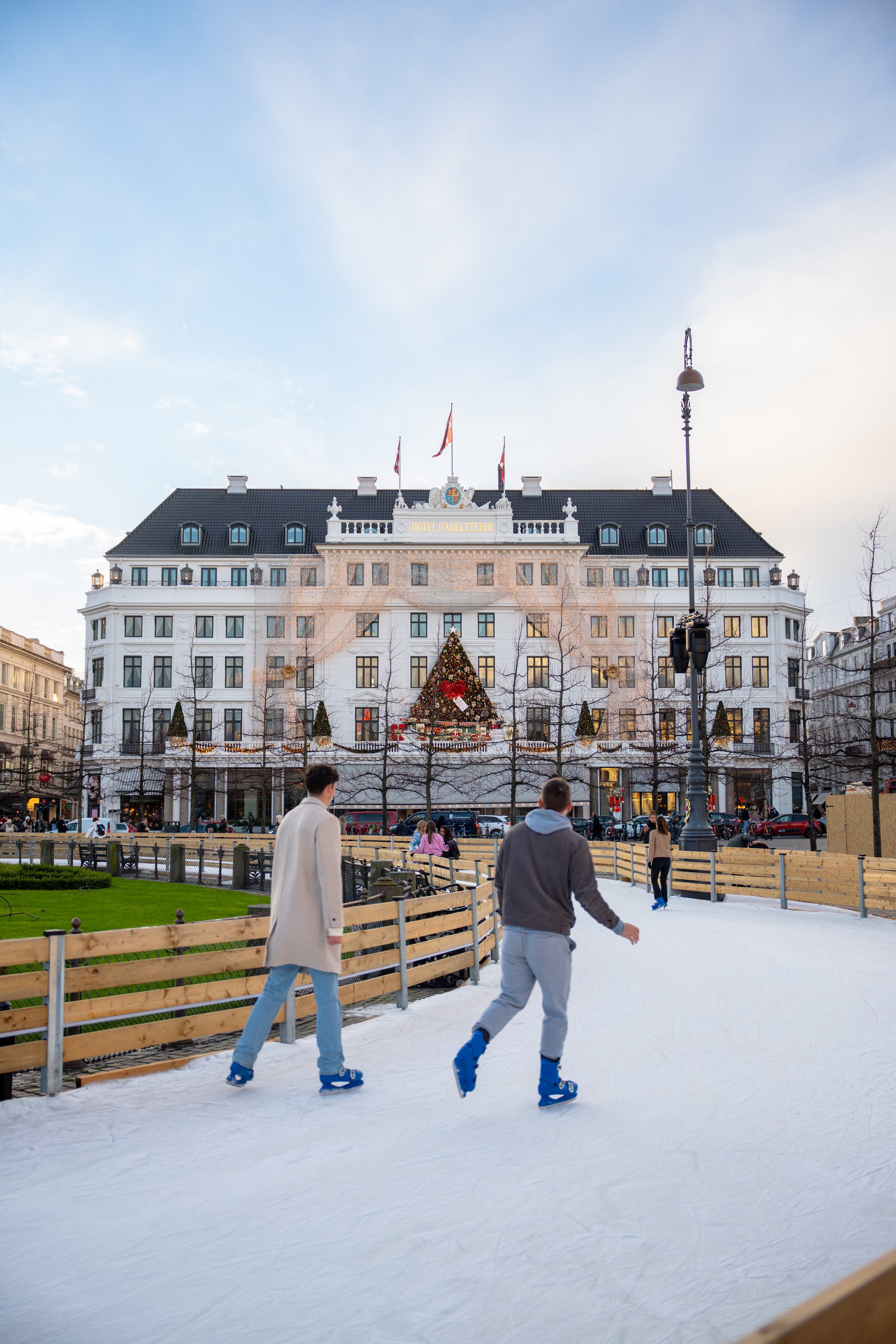 People skating on an outdoor ice rink in front of a large white hotel with a Christmas tree and Christmas decorations