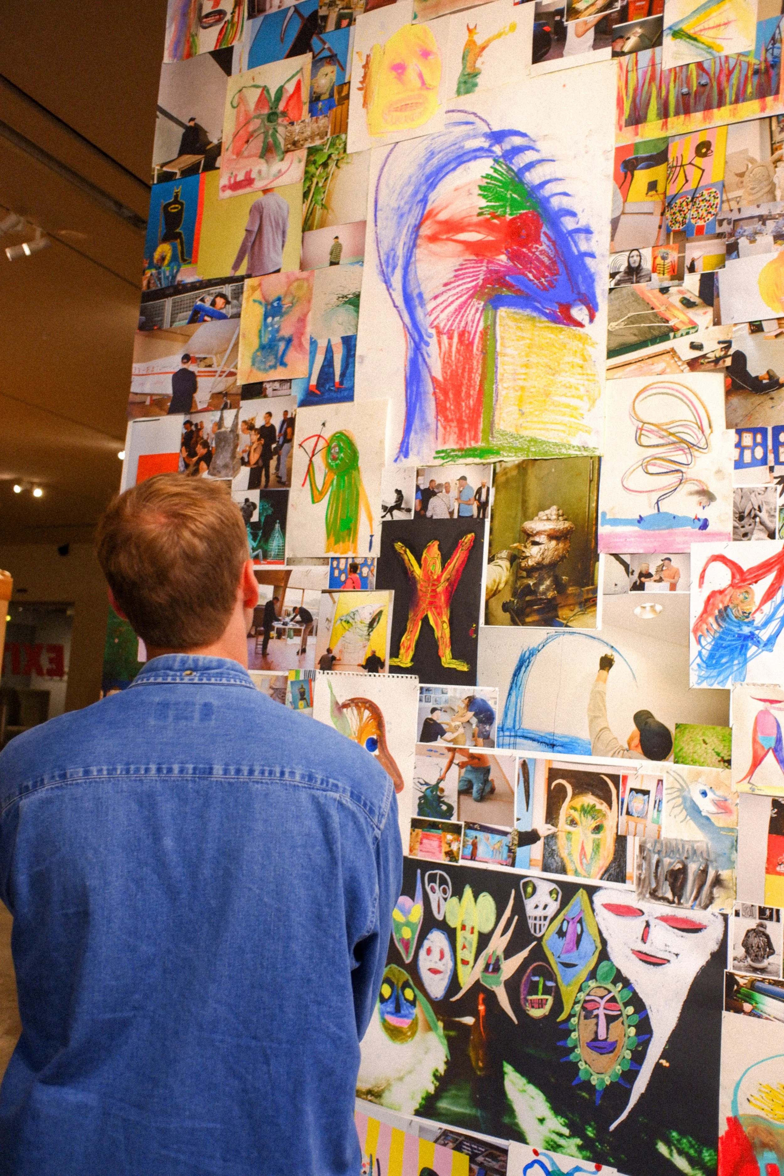 Person in blue denim jacket viewing a wall collage of colorful artworks, including drawings, paintings, and photographs, featuring abstract faces, figures, and patterns in a gallery or exhibition space.