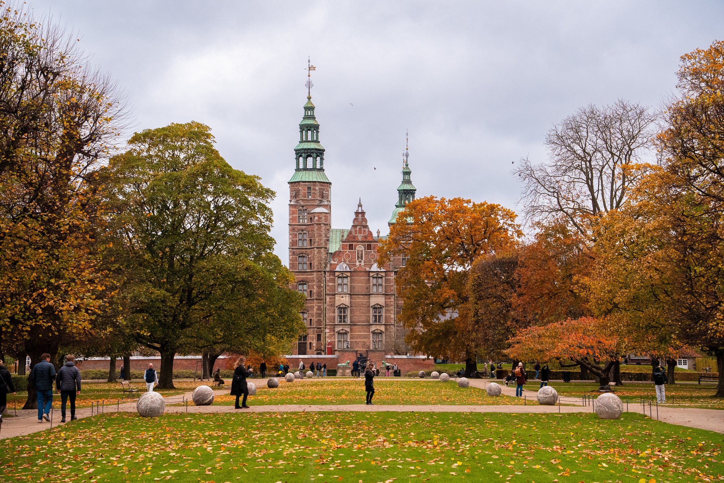 A historic castle-like building with green roofs and towers, surrounded by trees with autumn foliage, and people walking in a park with fallen leaves and large stone spheres in the foreground.