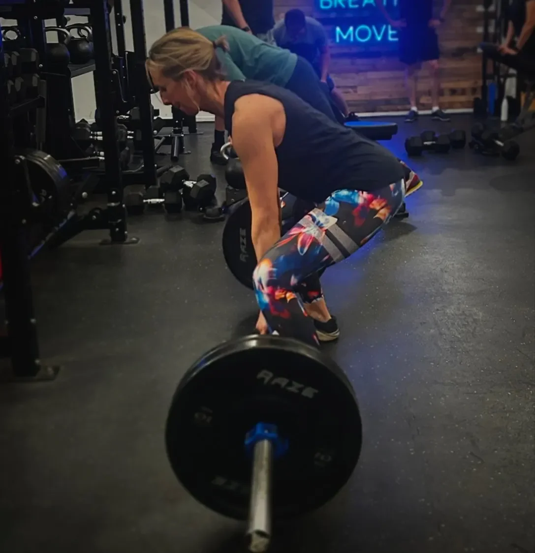 Woman performing a deadlift with a barbell in a gym, wearing colorful workout leggings and a black tank top.