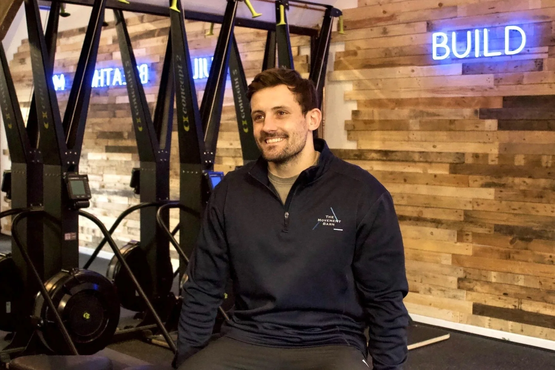A young man sitting on a bench in a neutral-colored room, wearing a gray polo shirt and beige pants, with a contemplative expression and a shadow cast on the wall behind him.