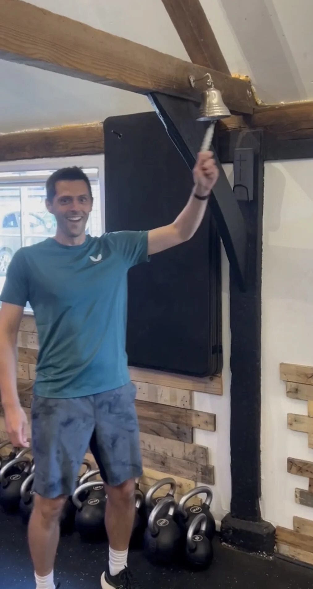 A man smiling and holding a bell with a workout rope attached, standing in a gym with kettlebells on the floor.