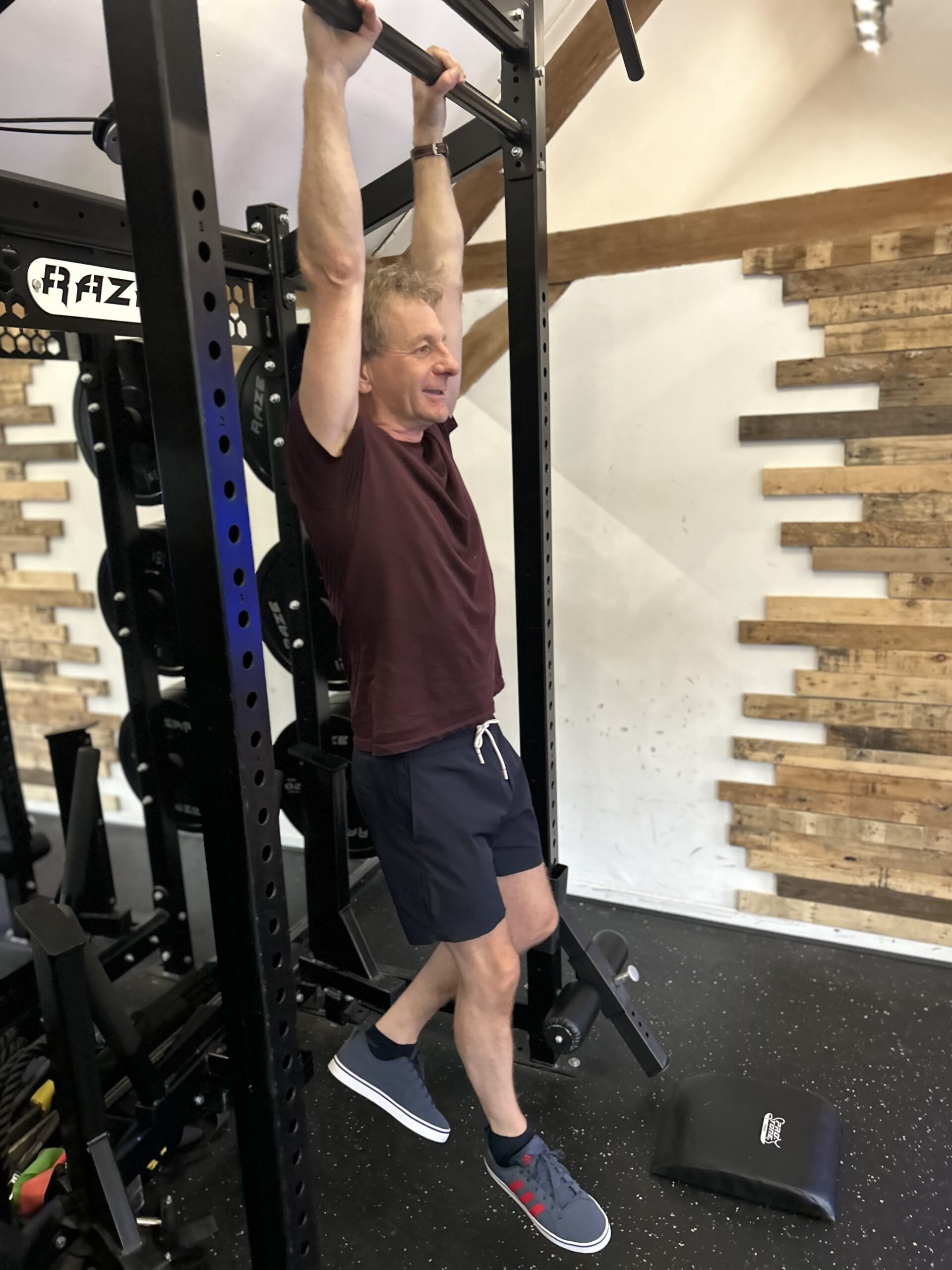 A man doing hanging exercise in a gym, gripping a pull-up bar with both hands, wearing a maroon T-shirt, navy shorts, and sneakers.